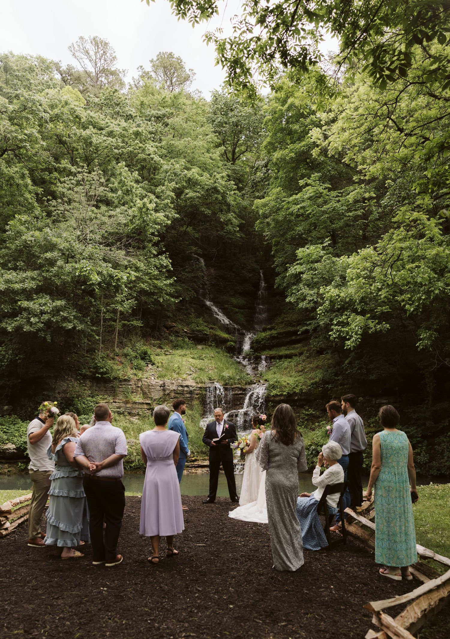 A wedding ceremony takes place in a lush forested area with a small gathering of guests and a waterfall backdrop.