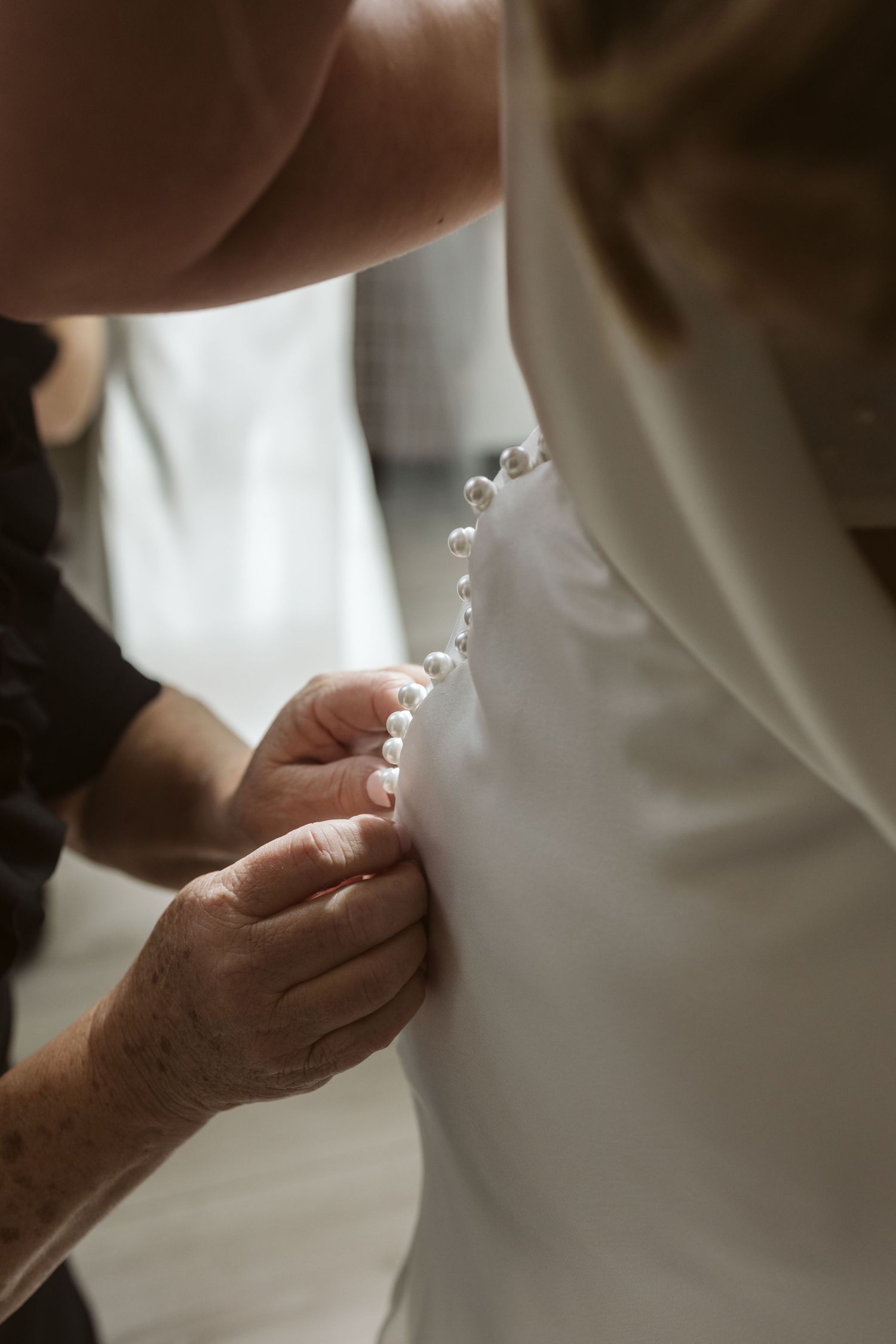 Close-up detail shot of hands fastening delicate pearl buttons on a white wedding dress.