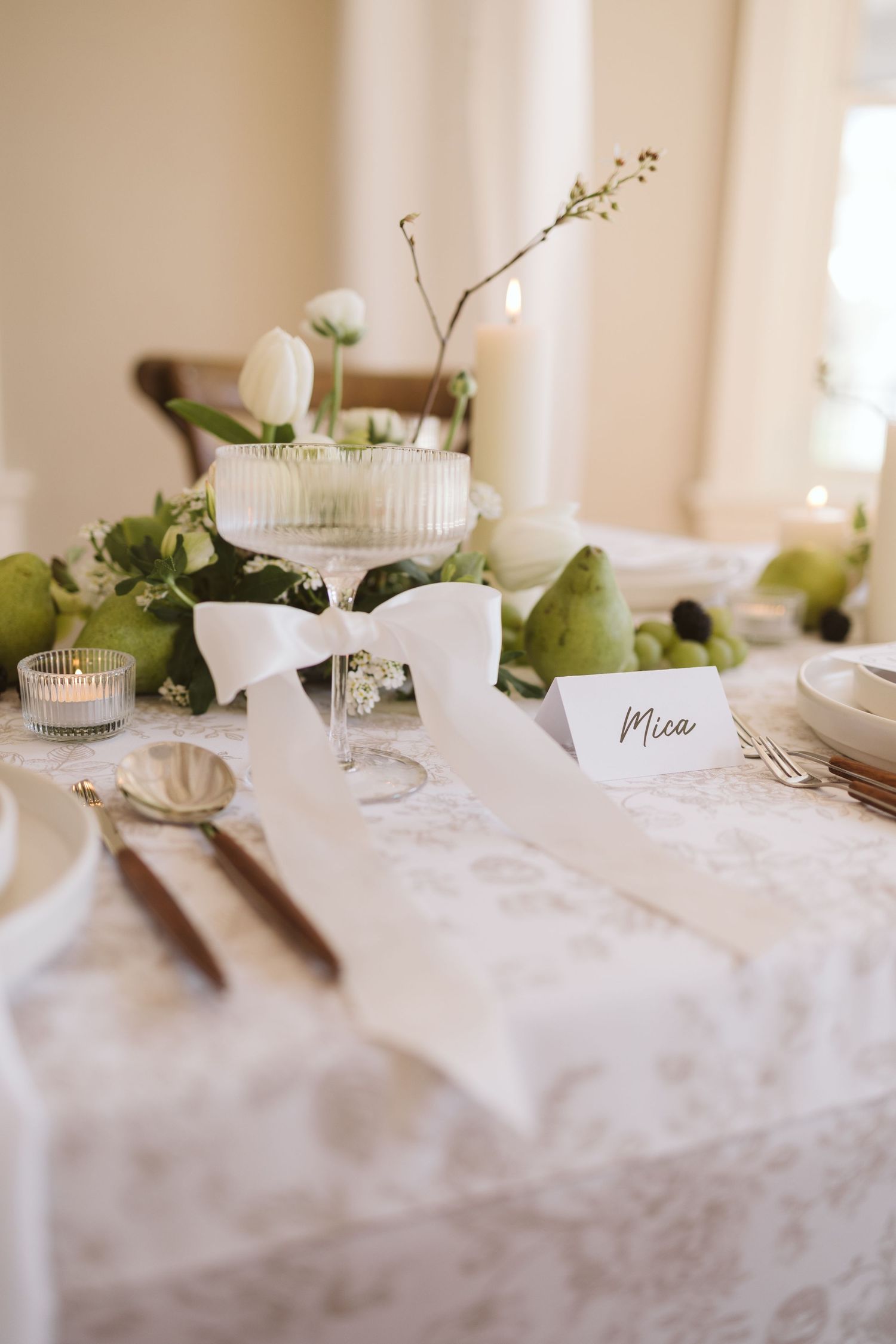 An elegant table setting with white florals, green pears, and a place card on a soft tablecloth in natural light.