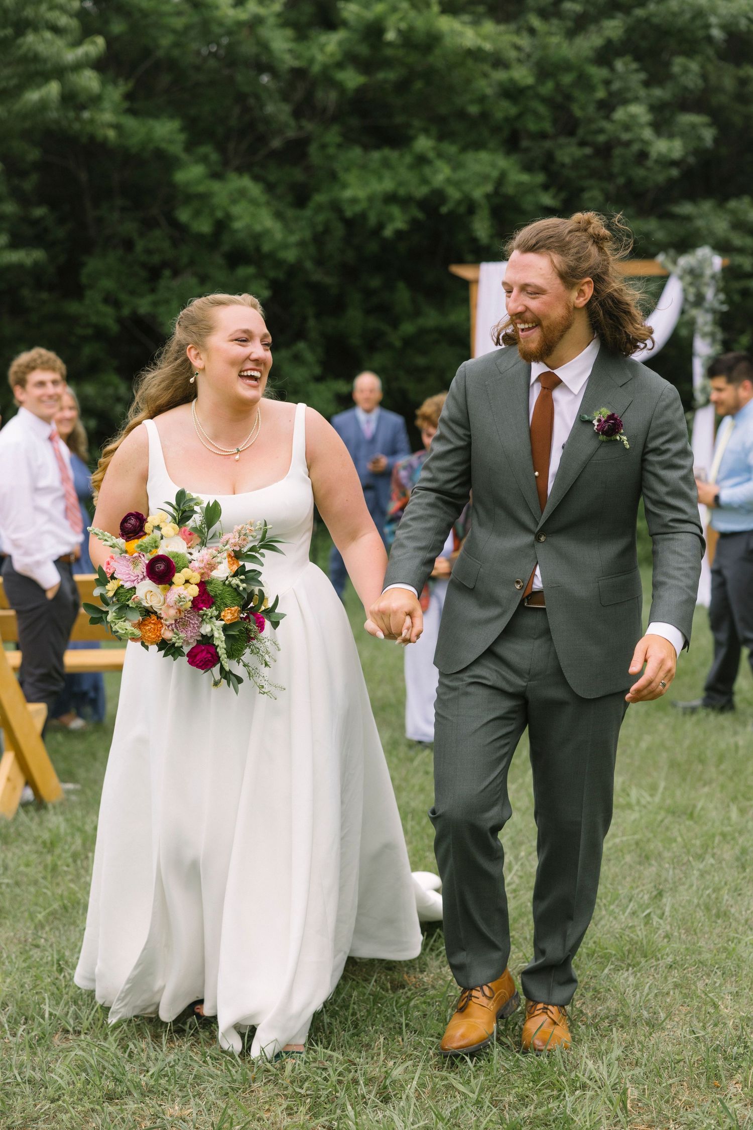 Wedding party dressed in white and dark suits walk playfully through a grassy outdoor venue during summer celebration.