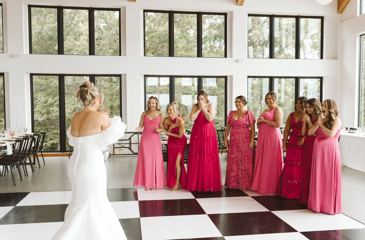 Wedding party stands on black and white checkered floor in bright indoor venue with pink gowns and bridal attire.
