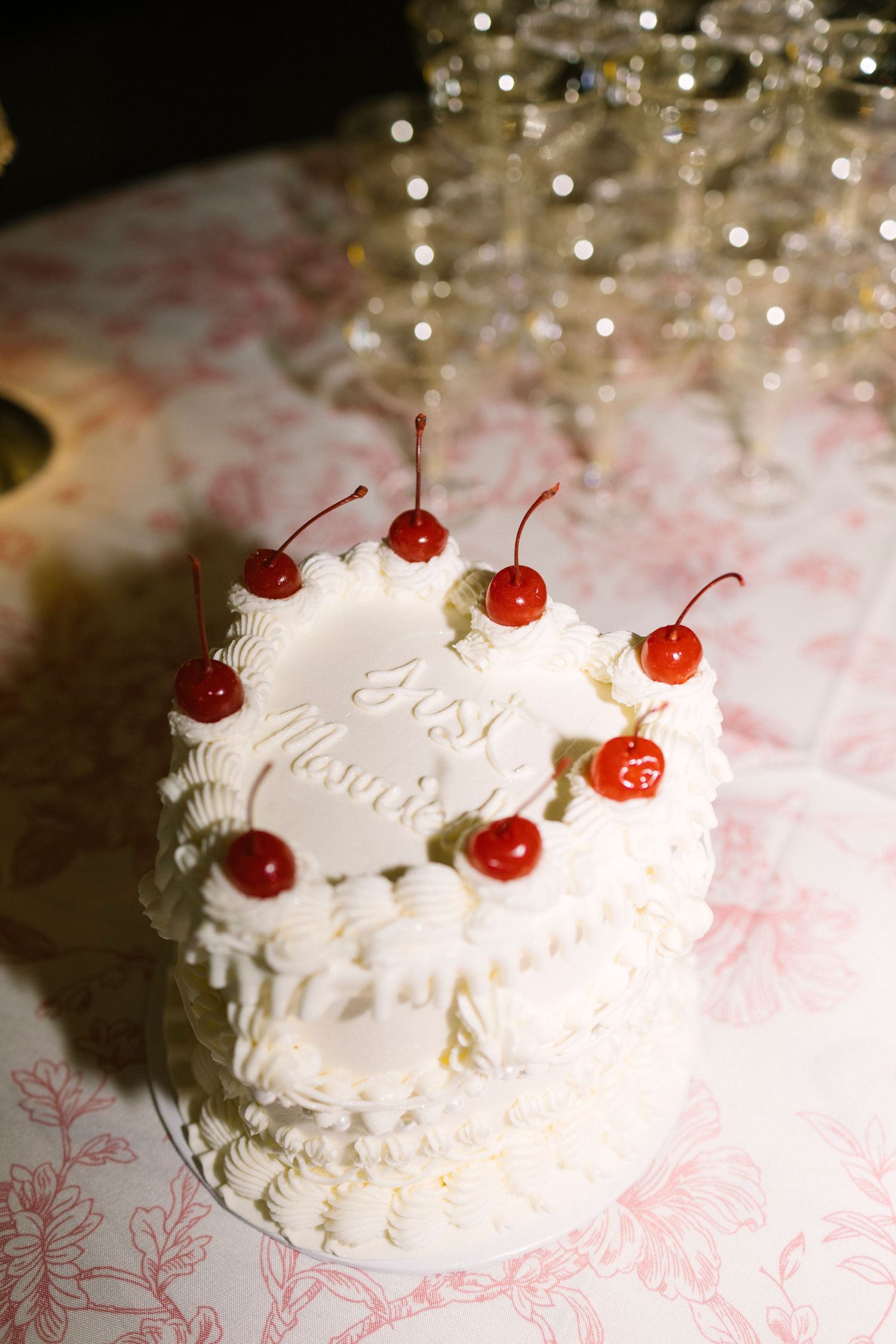 Elegant white wedding cake decorated with bright red maraschino cherries and delicate piping on pink floral tablecloth.