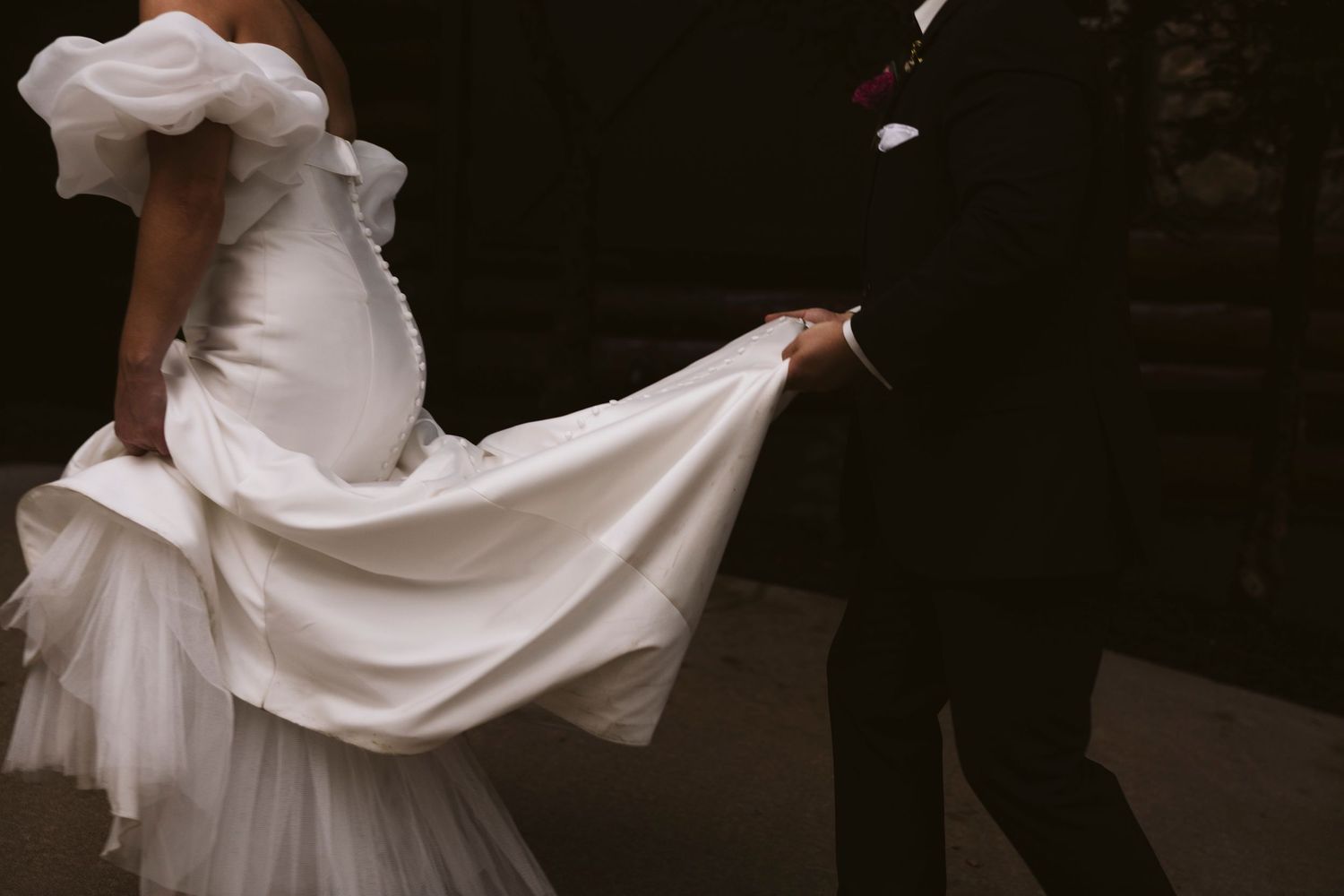 A flowing white wedding dress being held up while walking beside someone in a dark suit.