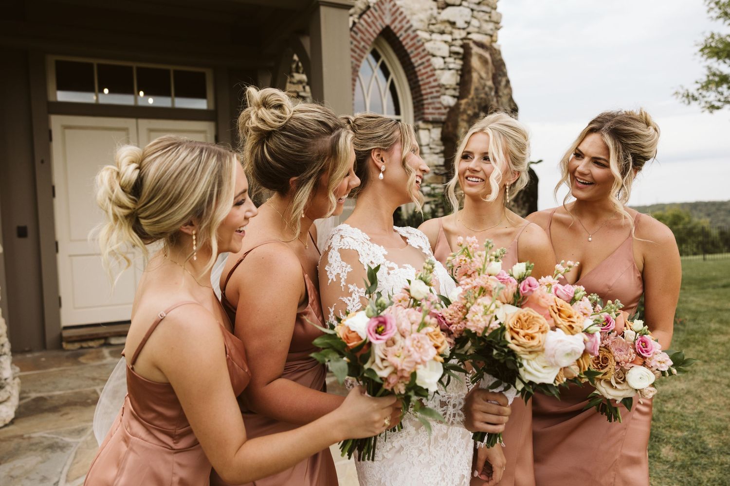 Candid moment of bridal party sharing laughs while holding peach and white bouquets.
