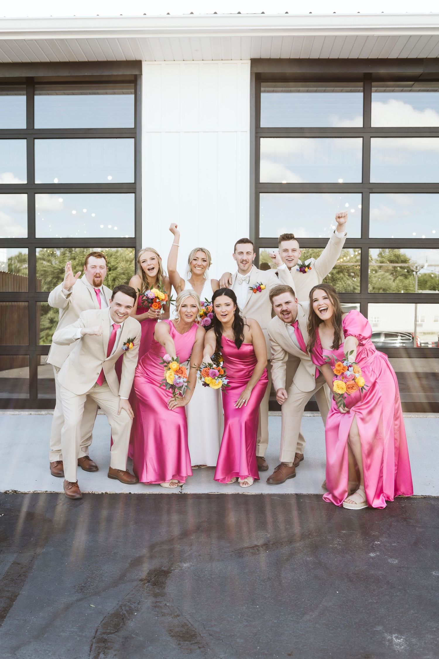 Wedding party poses together wearing tan suits and pink dresses with colorful bouquets against modern glass doors.