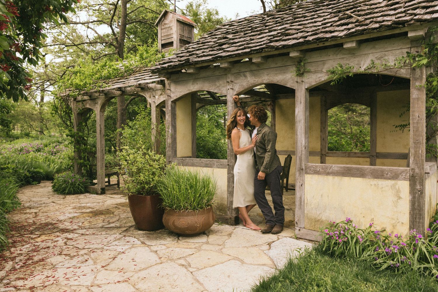 A couple standing under a rustic covered porch with stone floors and potted plants.