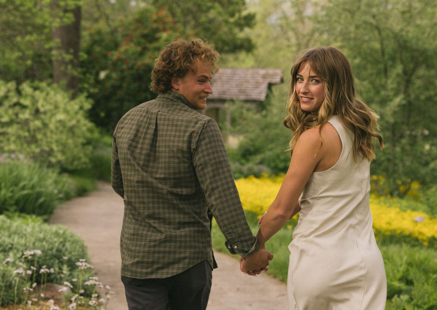 Couple holding hands and walking down a path surrounded by lush greenery and yellow wildflowers on a sunny day.