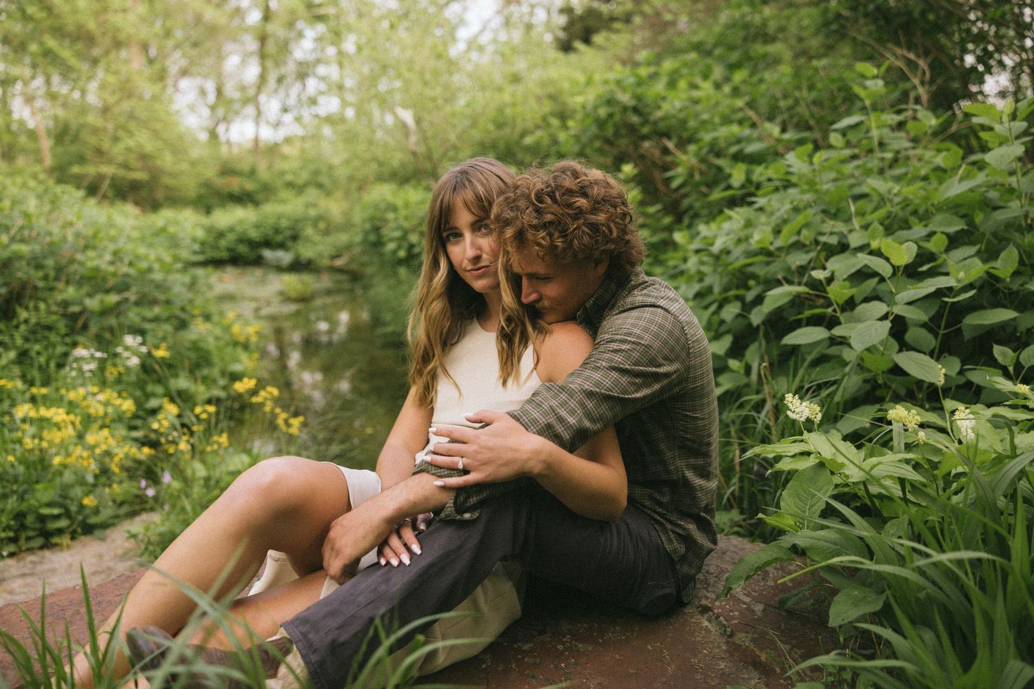 A couple sits together in a lush garden setting sharing an intimate embrace on a summer afternoon.