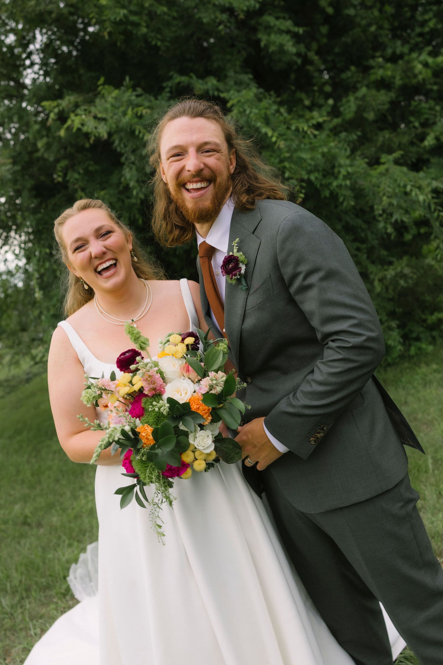 A candid moment of laughter shared between newlyweds with a colorful bouquet in an outdoor setting.