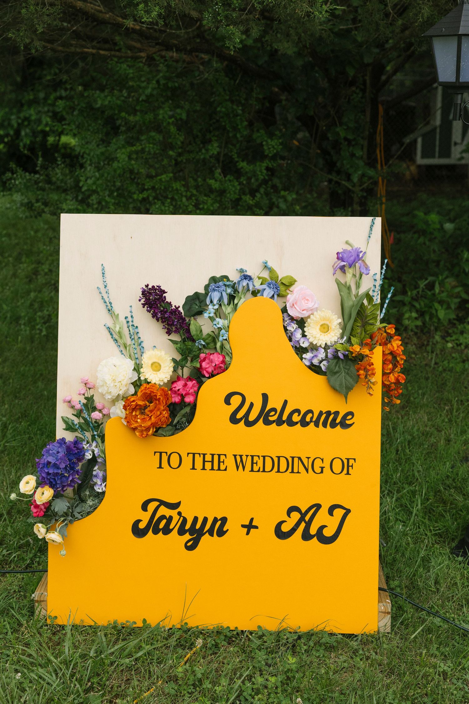 Yellow welcome wedding sign decorated with colorful fresh flowers and elegant lettering displayed on grass outdoors.