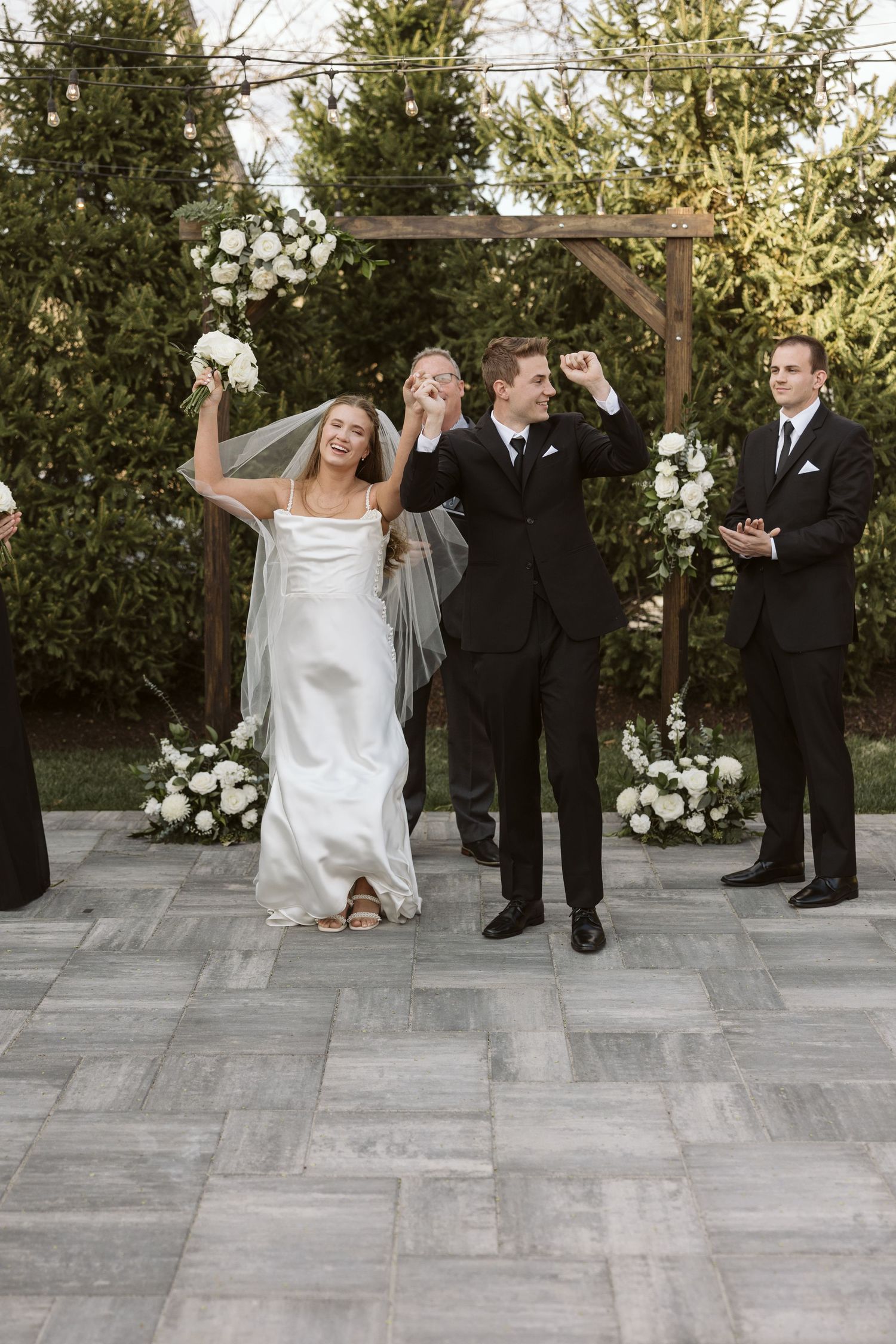 A playful group shot of the wedding party standing on gray stone tiles with greenery in the background.