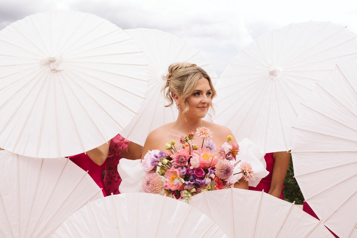 A bride in white dress holds pink and purple bouquet surrounded by white paper parasols on her wedding day.