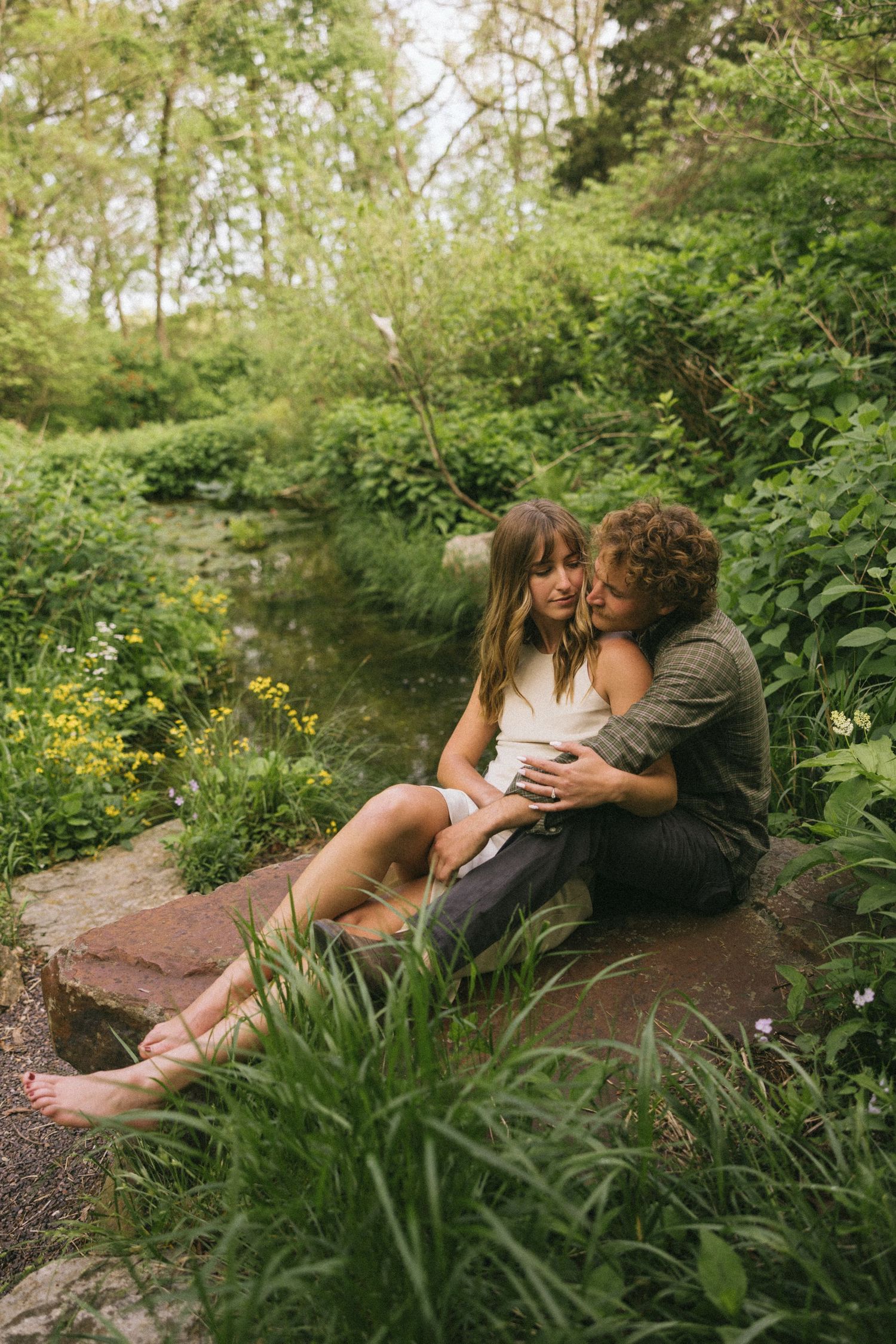 A couple sits intimately by a peaceful stream surrounded by lush greenery and yellow wildflowers on a summer day.