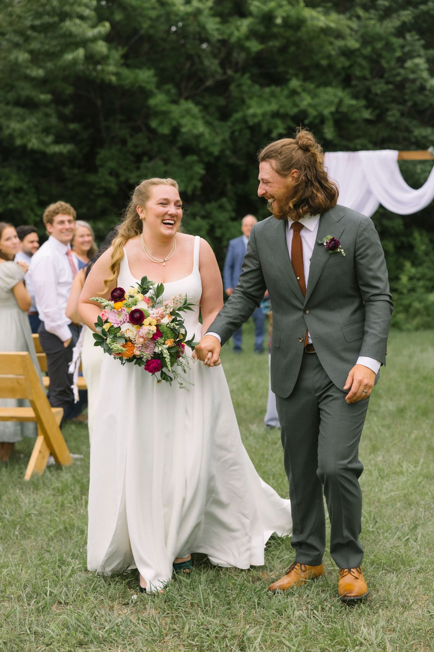 A joyous outdoor wedding ceremony with couple walking and laughing together among trees and guests.