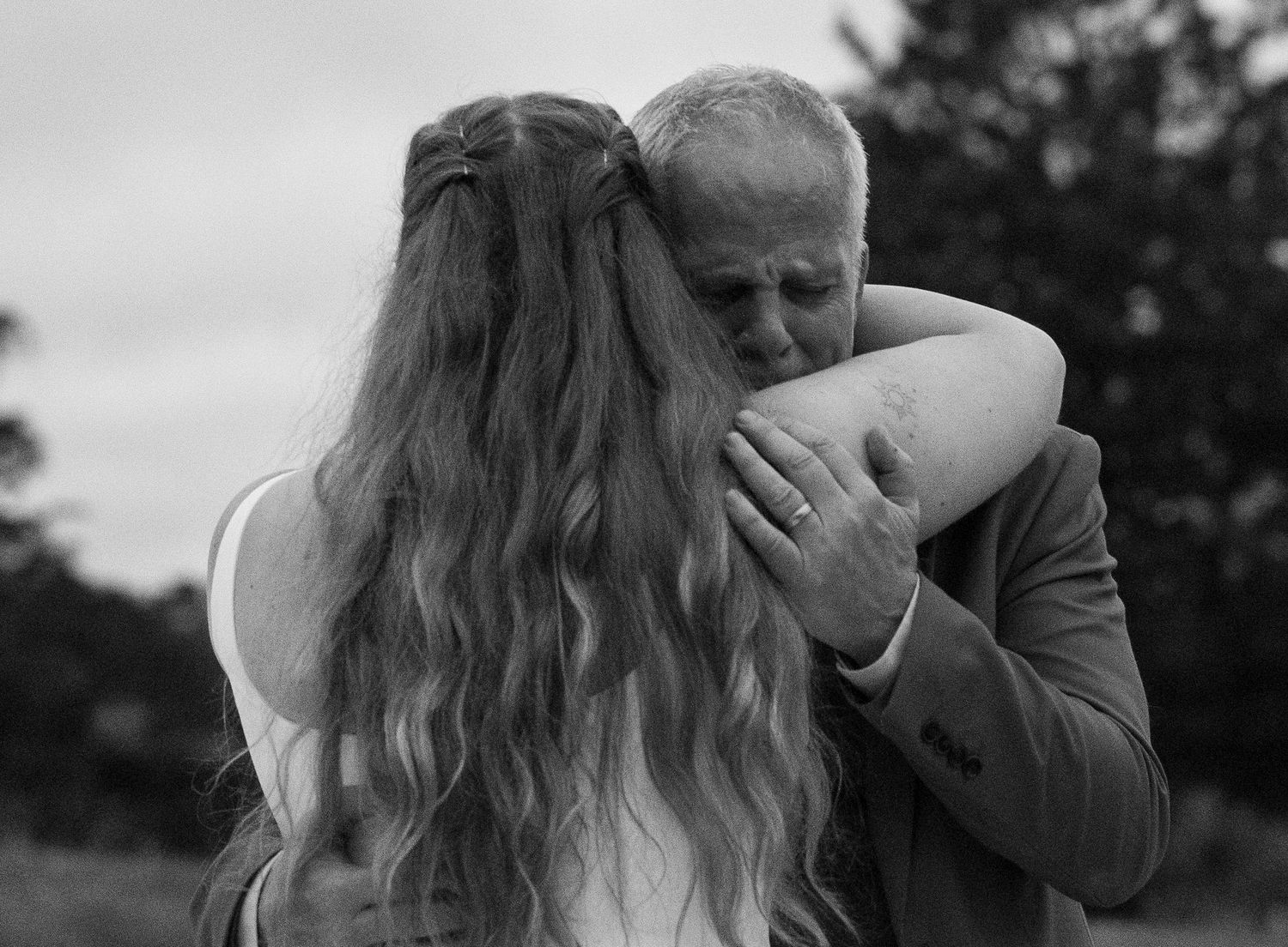 Black and white emotional moment captured between an elderly man and a woman with long wavy hair embracing at outdoor event.