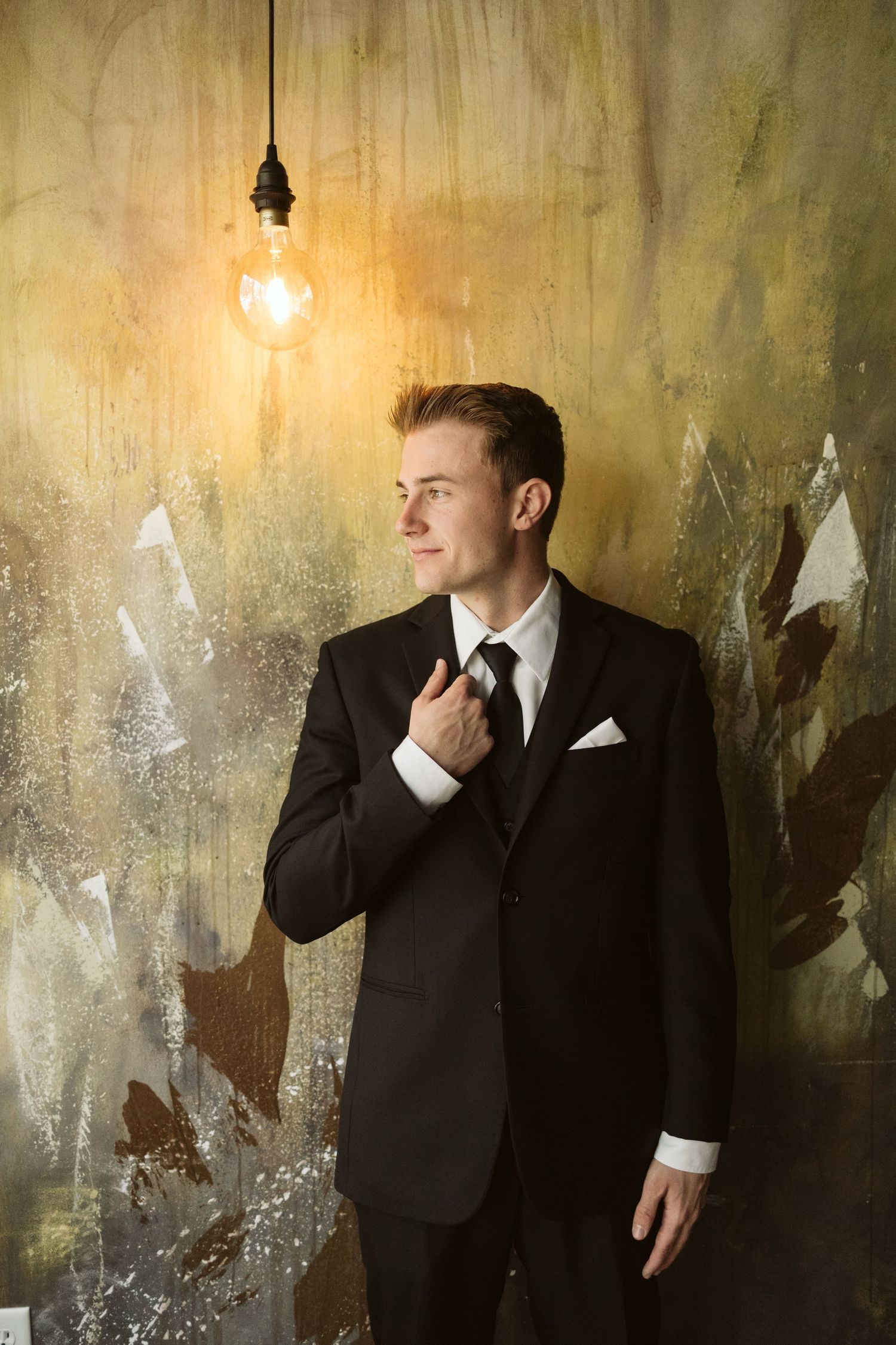 A groom in a classic black tuxedo stands against a textured wall with a glowing vintage light bulb creating moody ambiance.