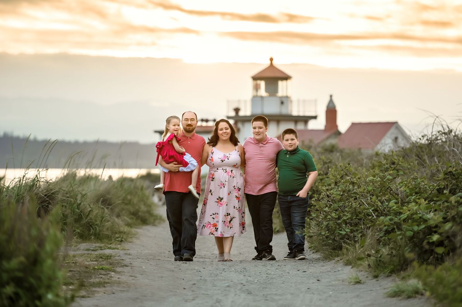 Family walking on path at sunset with lighthouse and buildings in background.