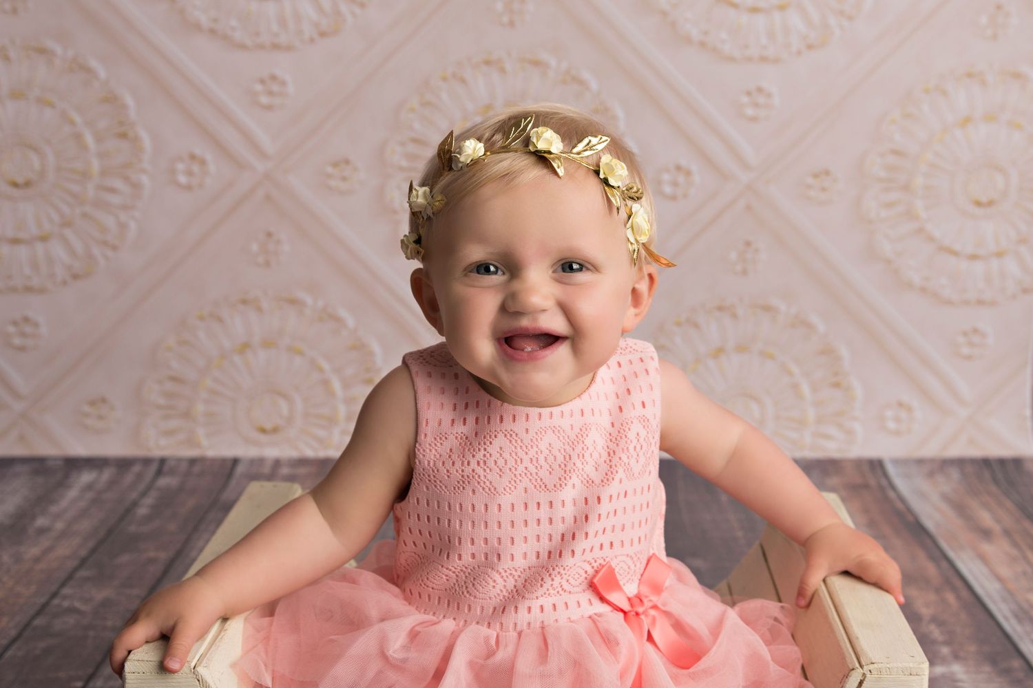 A happy baby wearing a pink tulle dress and floral headband sits on a wooden floor against a patterned wall.