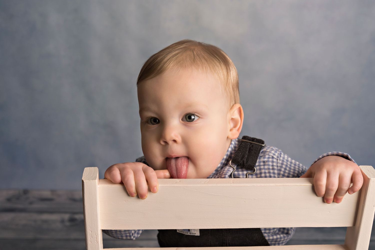 A small child in a checkered shirt sticks out their tongue while peeking over a white wooden chair against a gray backdrop.