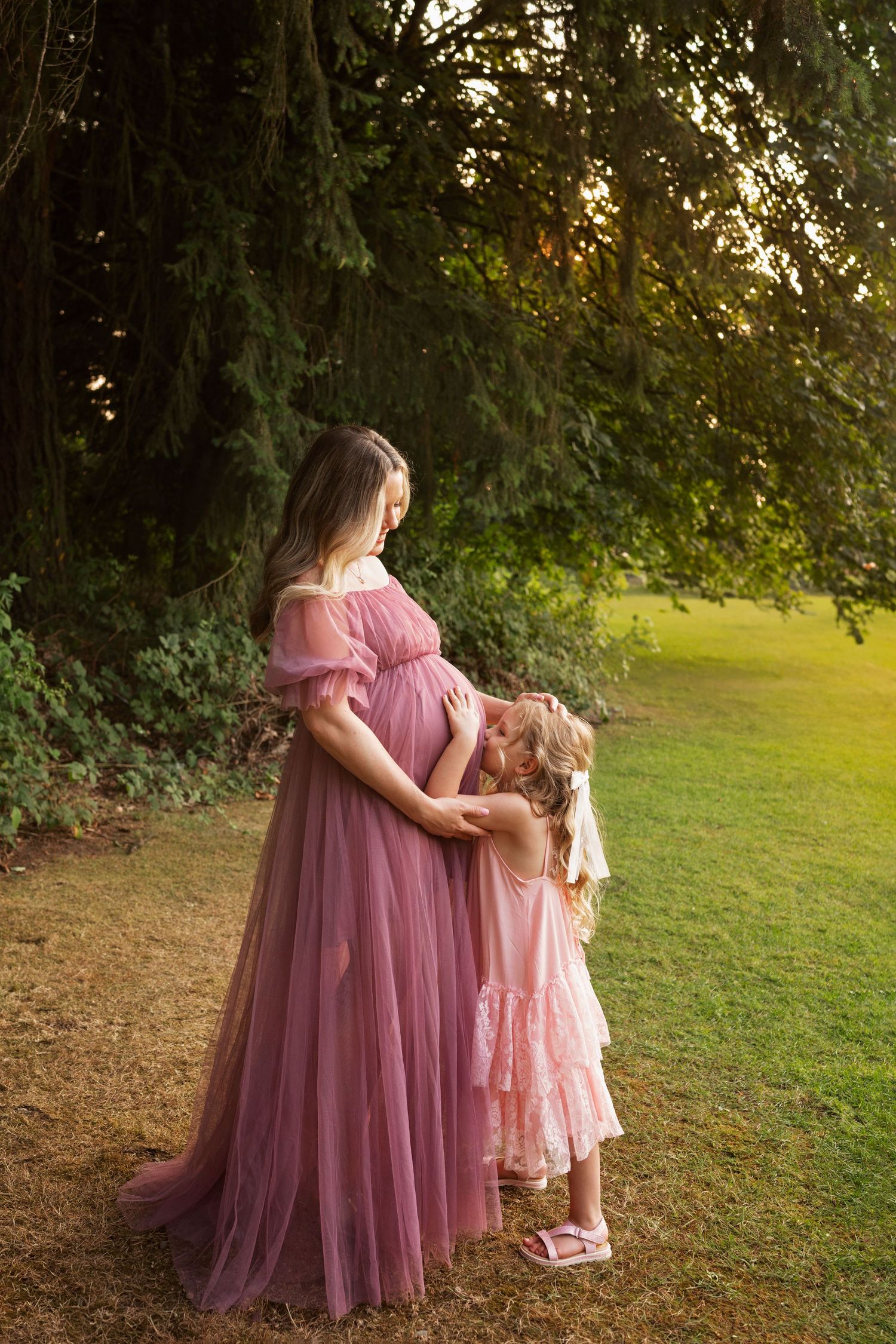 Mother in mauve gown and daughter in pink dress sharing tender moment outdoors at sunset.