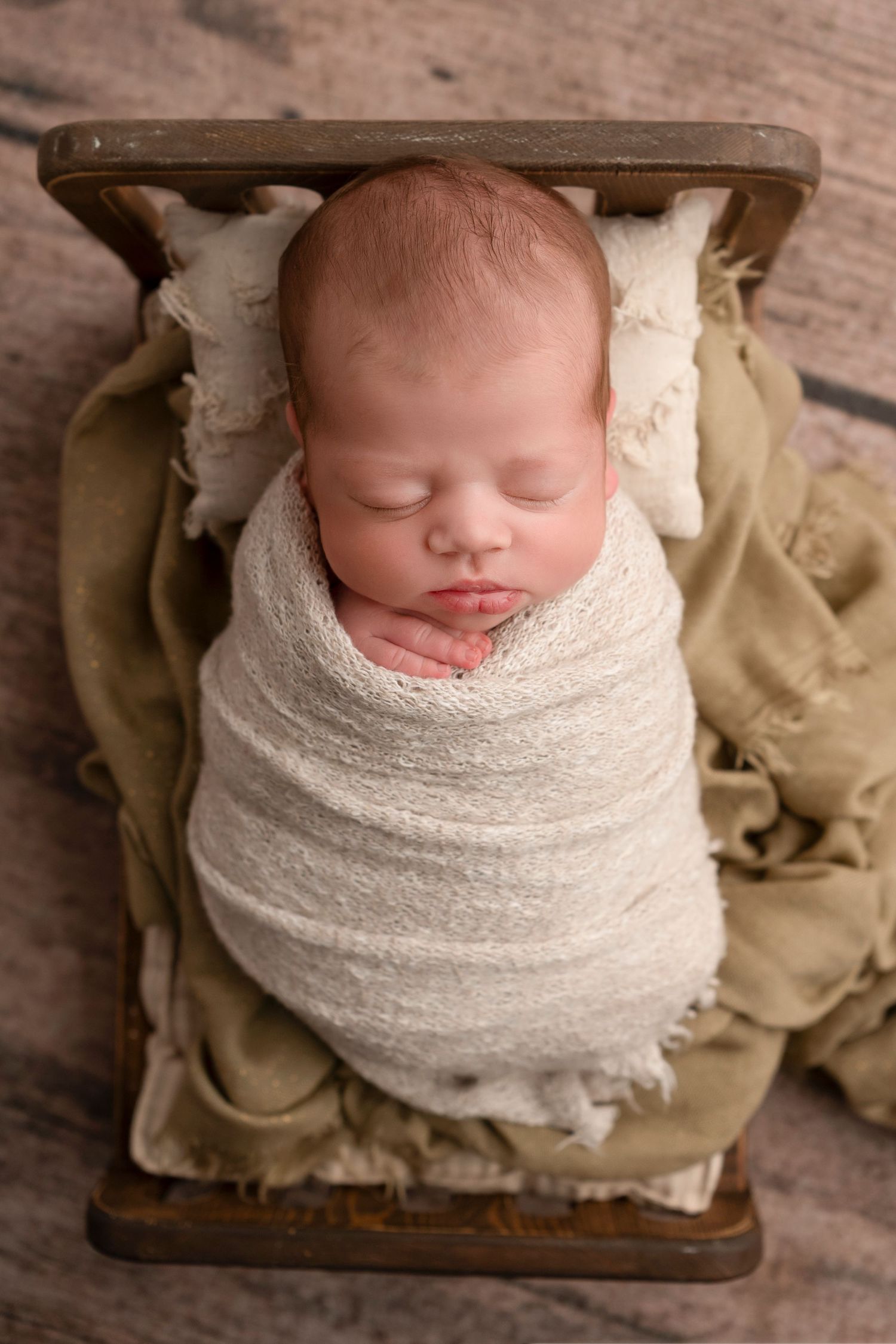 Newborn sleeping peacefully wrapped in white swaddle on vintage wooden prop.