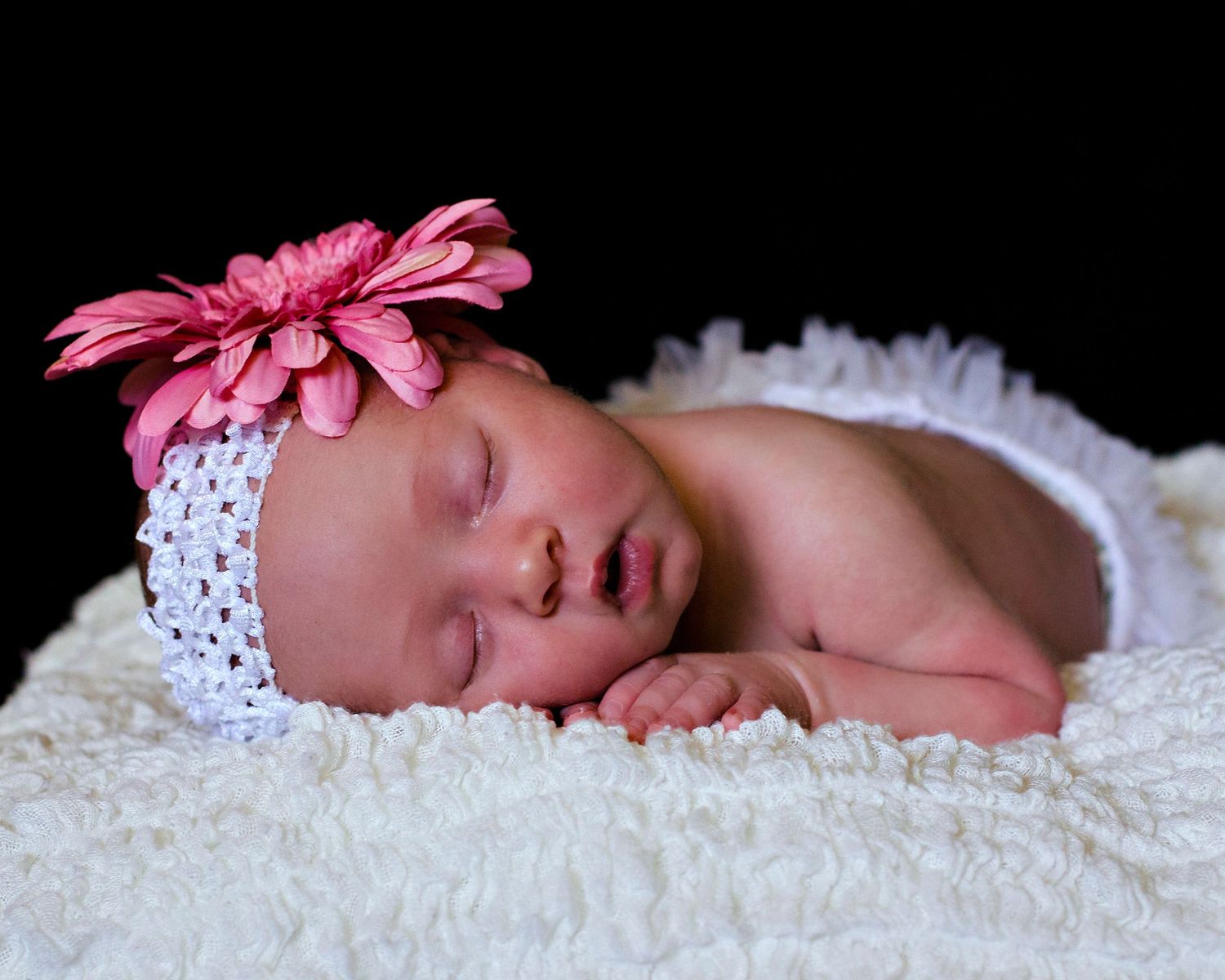 Peaceful sleeping baby wearing a white headband with pink flower lies on soft white blanket against black background.