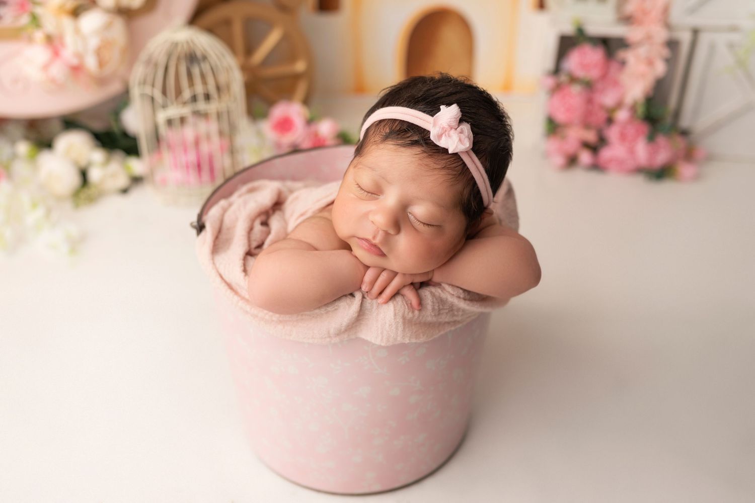 Sleeping newborn wearing pink headband rests peacefully in pink bucket with floral decorations in background.
