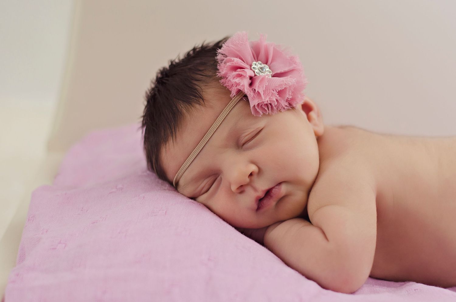 A sleeping newborn baby with soft pink floral headband lying on white blankets during a peaceful photoshoot.