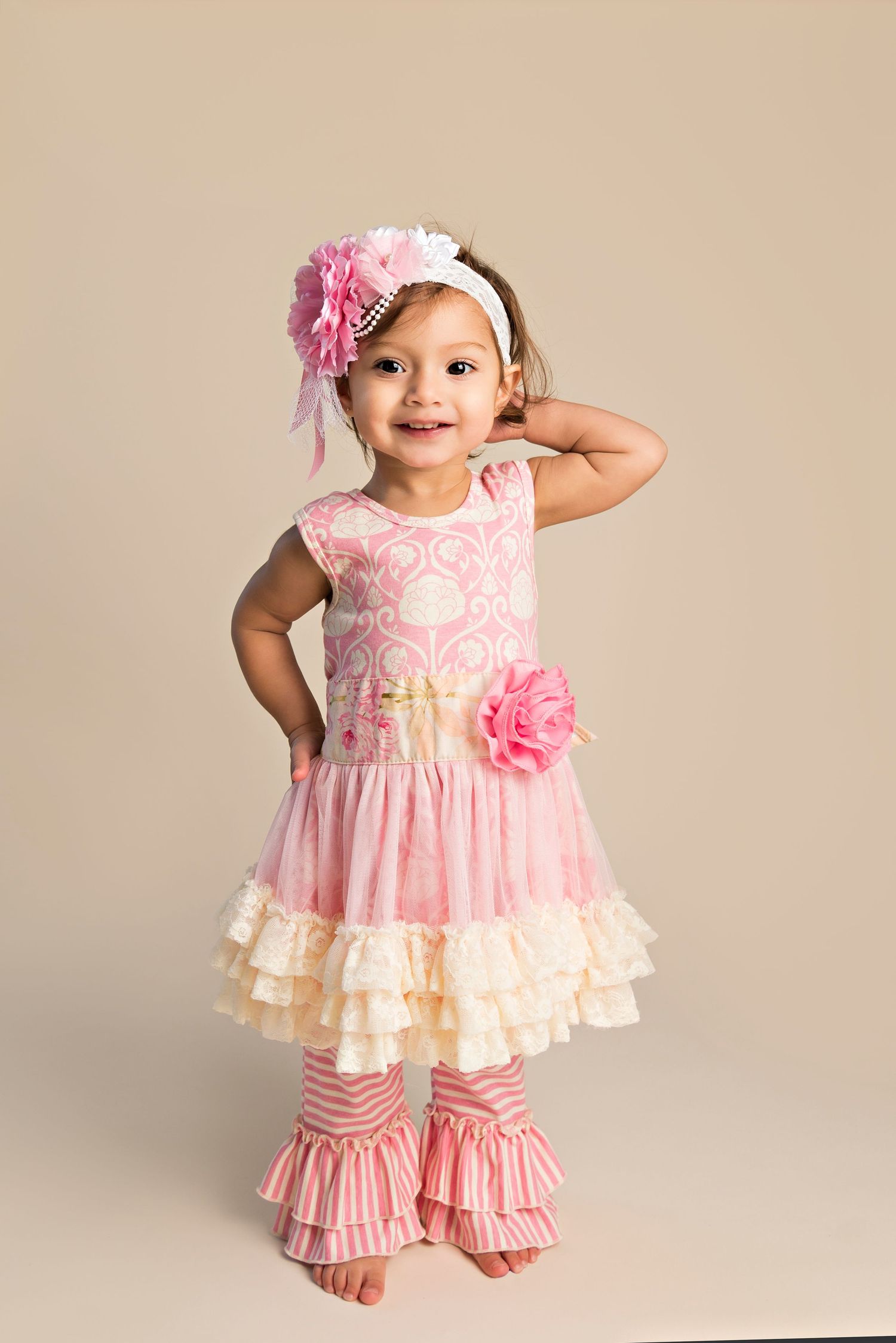 A small child wearing a pink and cream ruffled dress with matching striped pants and floral headband poses against beige background.