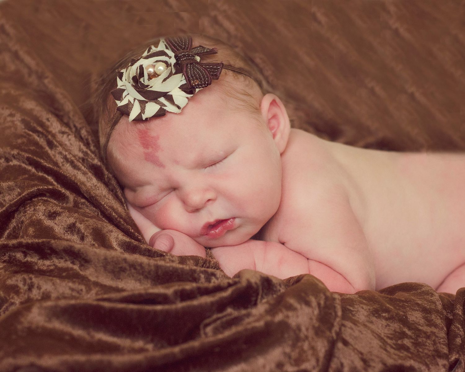Newborn baby sleeping peacefully on soft brown blanket with decorative floral headband.