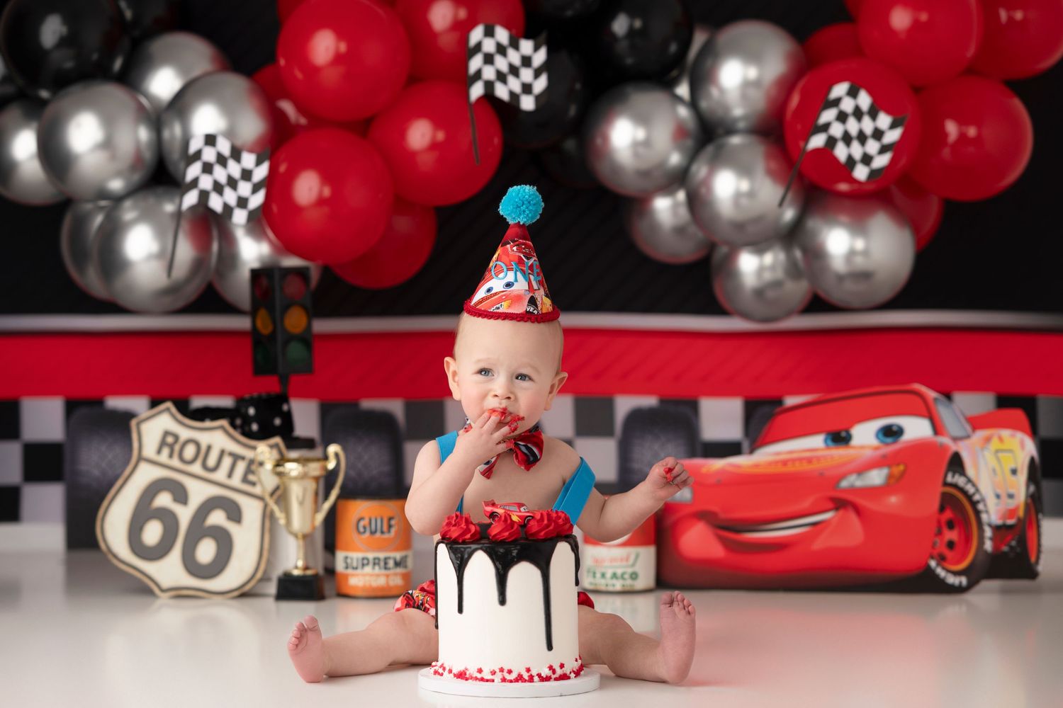 Baby with party hat sitting by Cars themed birthday cake with red and silver balloon backdrop.
