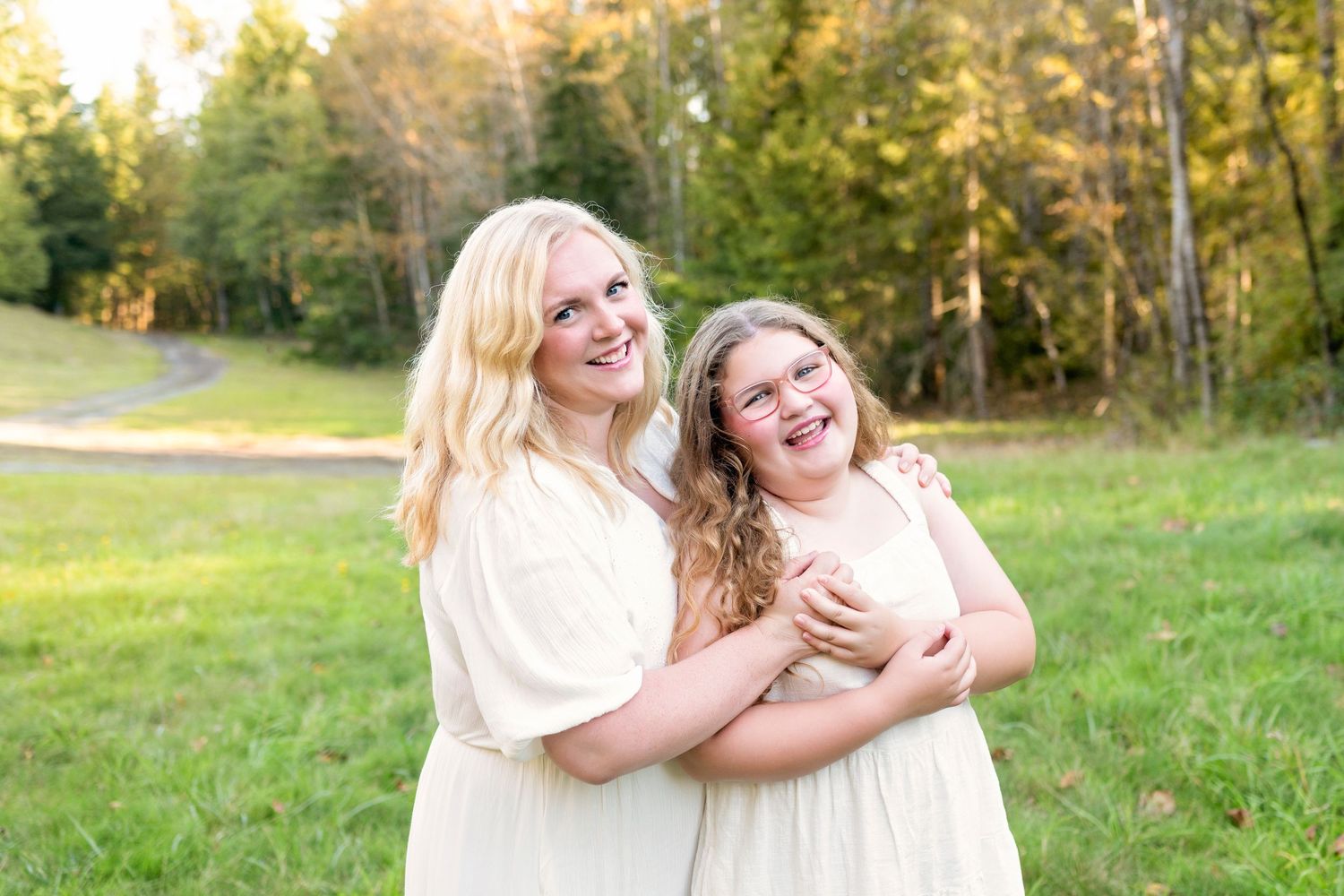 Mother and daughter embracing while wearing matching white dresses in a sunny field.