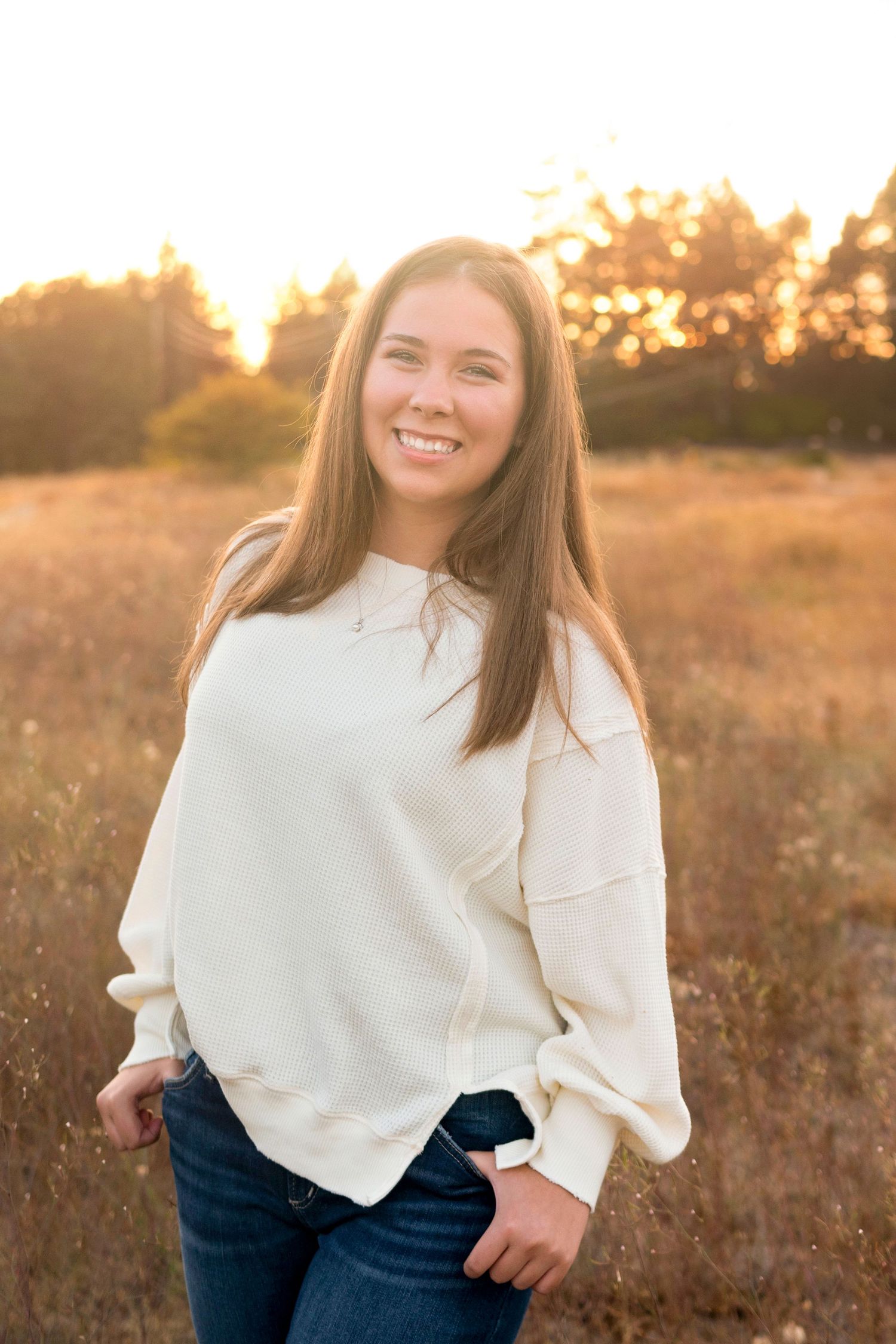 Woman in white sweater smiling in golden sunlit field during sunset.