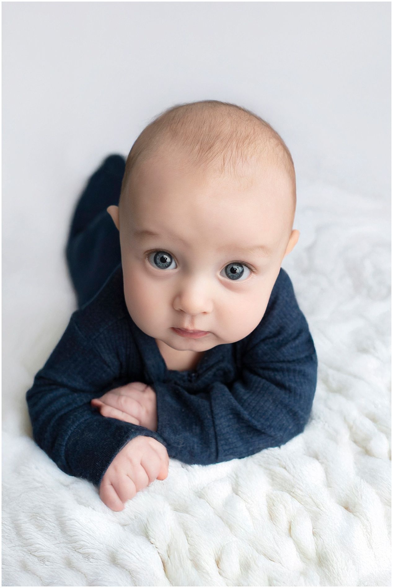 Baby in navy sweater lying on white textured blanket during professional photo session.