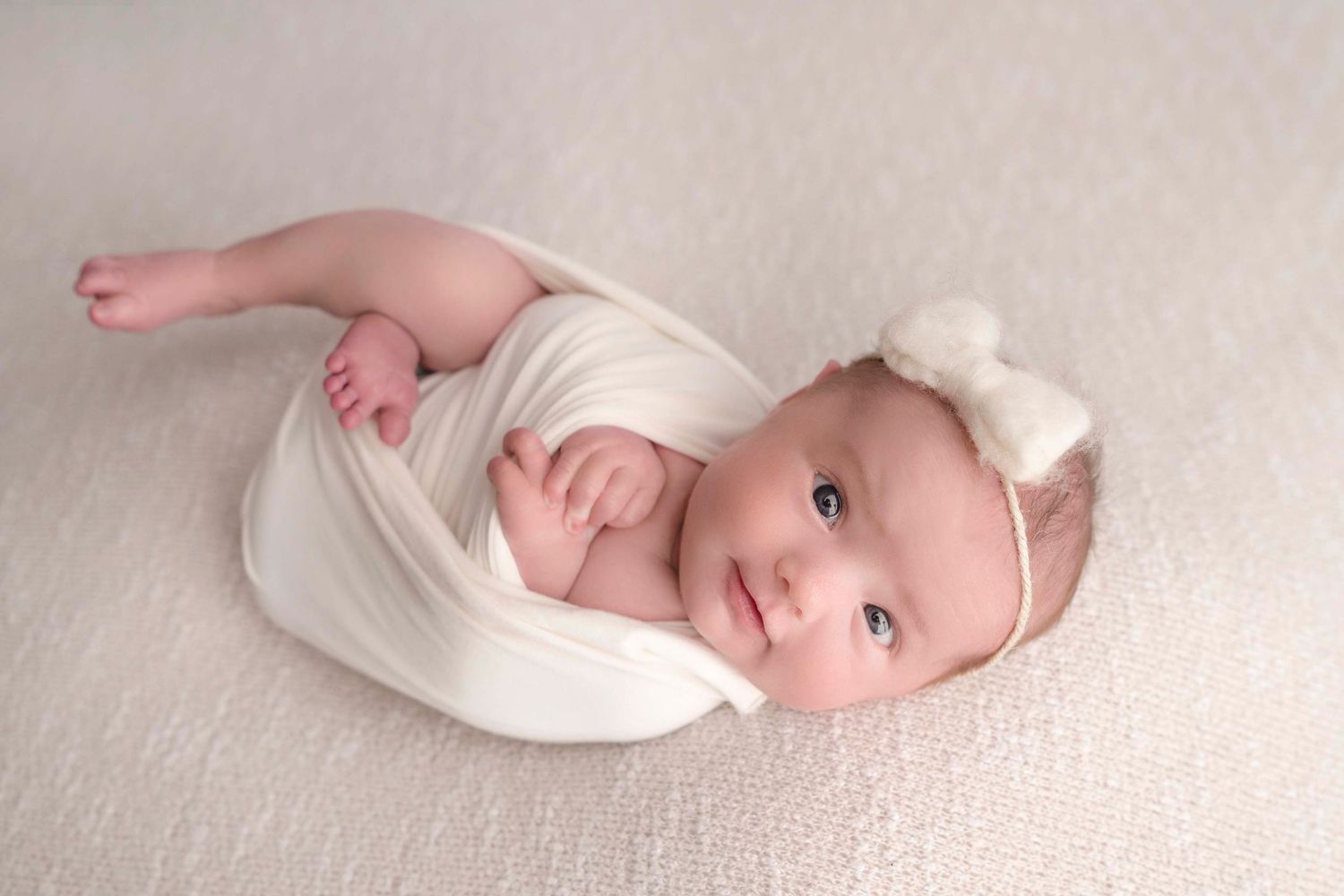 A newborn baby wrapped in white fabric lies on a cream-colored background with a white bow headband.