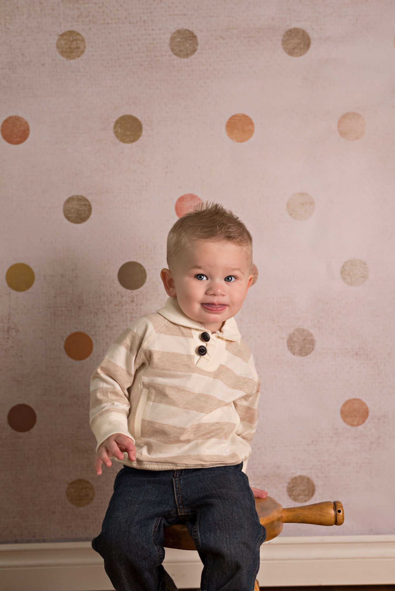 A baby in cream colored sweater and jeans sits against polka dot wall paper background showing different expressions.