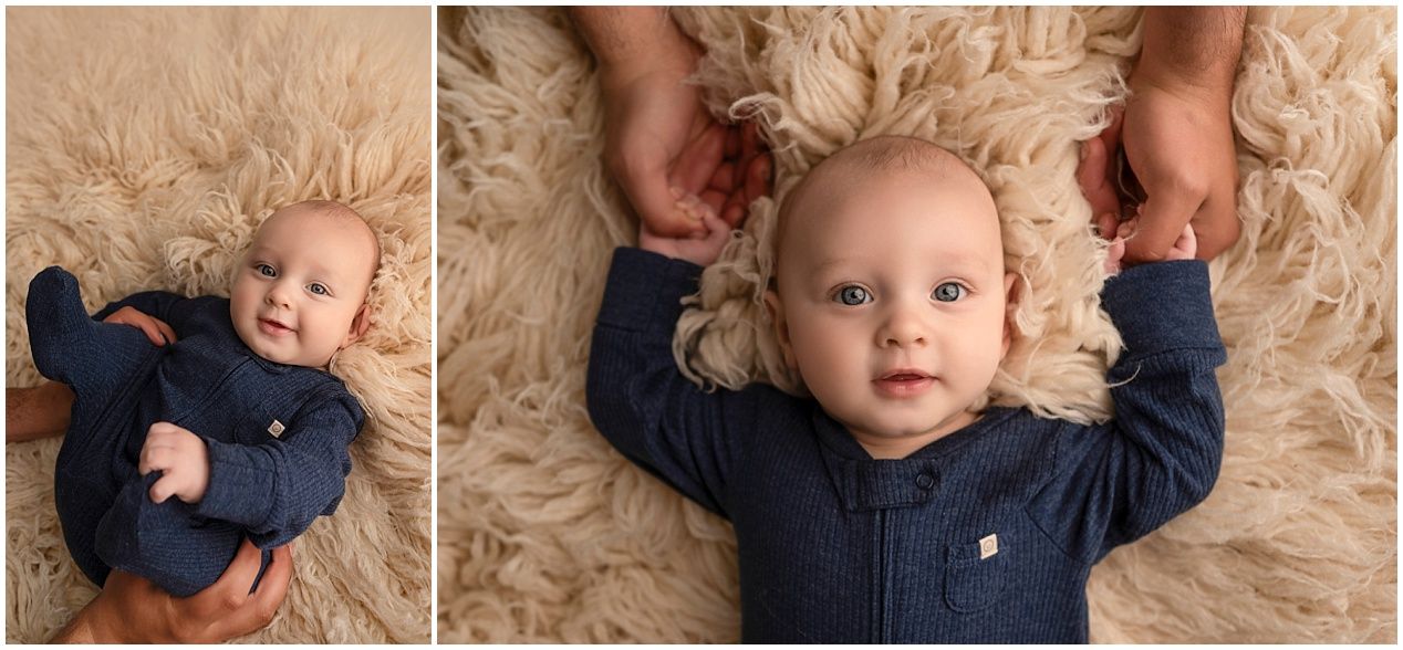 Baby wearing navy outfit smiles while lying on beige fuzzy rug during professional photoshoot.