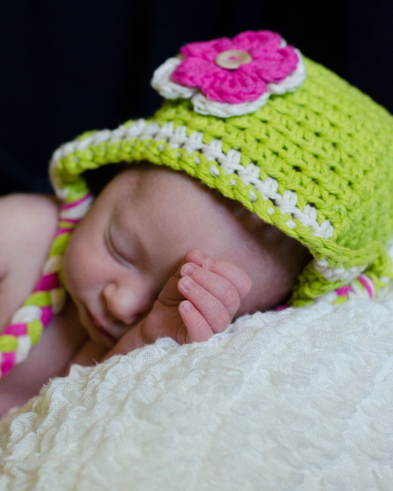 A baby wearing a bright yellow crocheted hat with pink flower trim sleeps peacefully on white textured blanket.