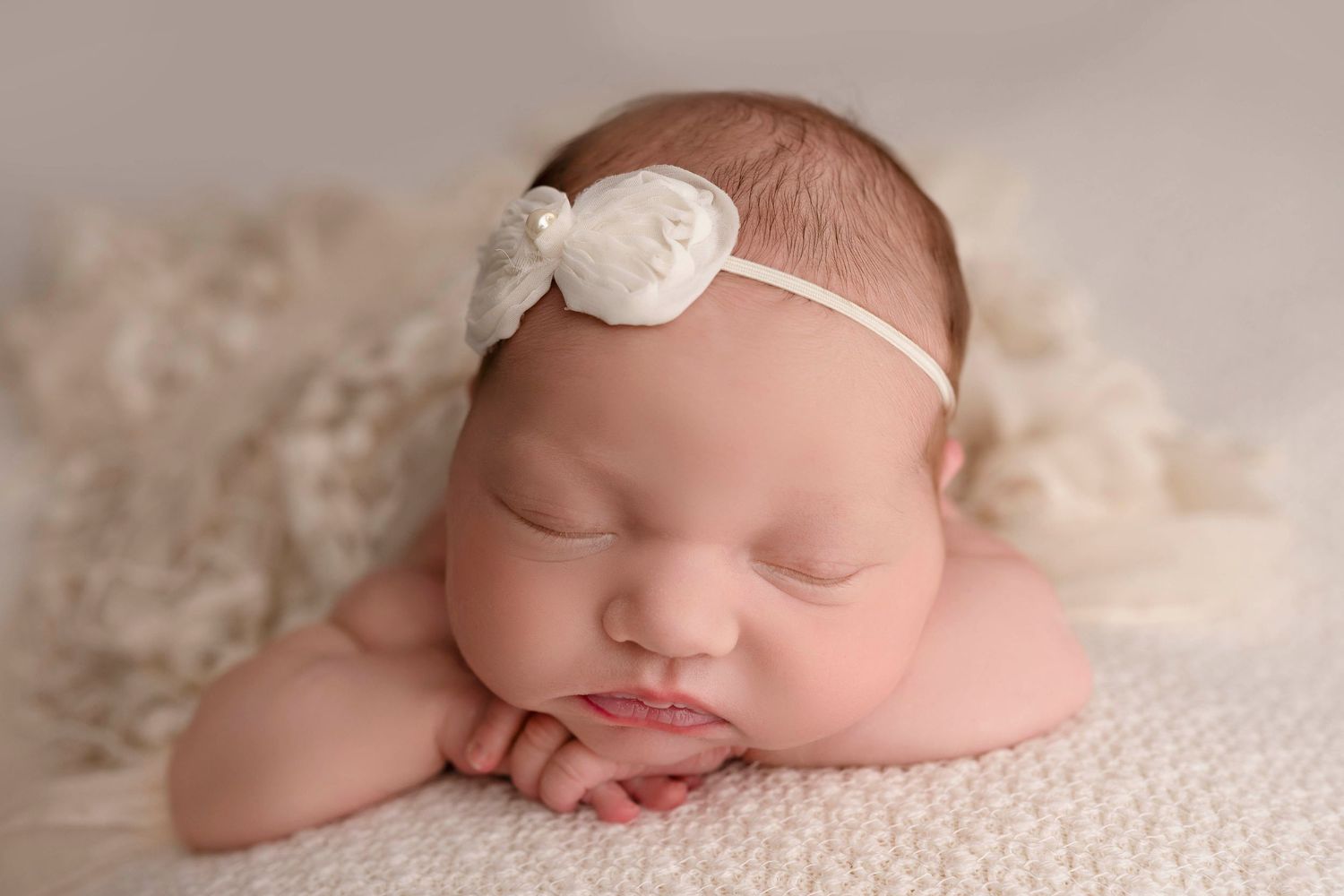 Sleeping newborn wearing delicate white headband rests peacefully on soft cream-colored blanket during photo session.