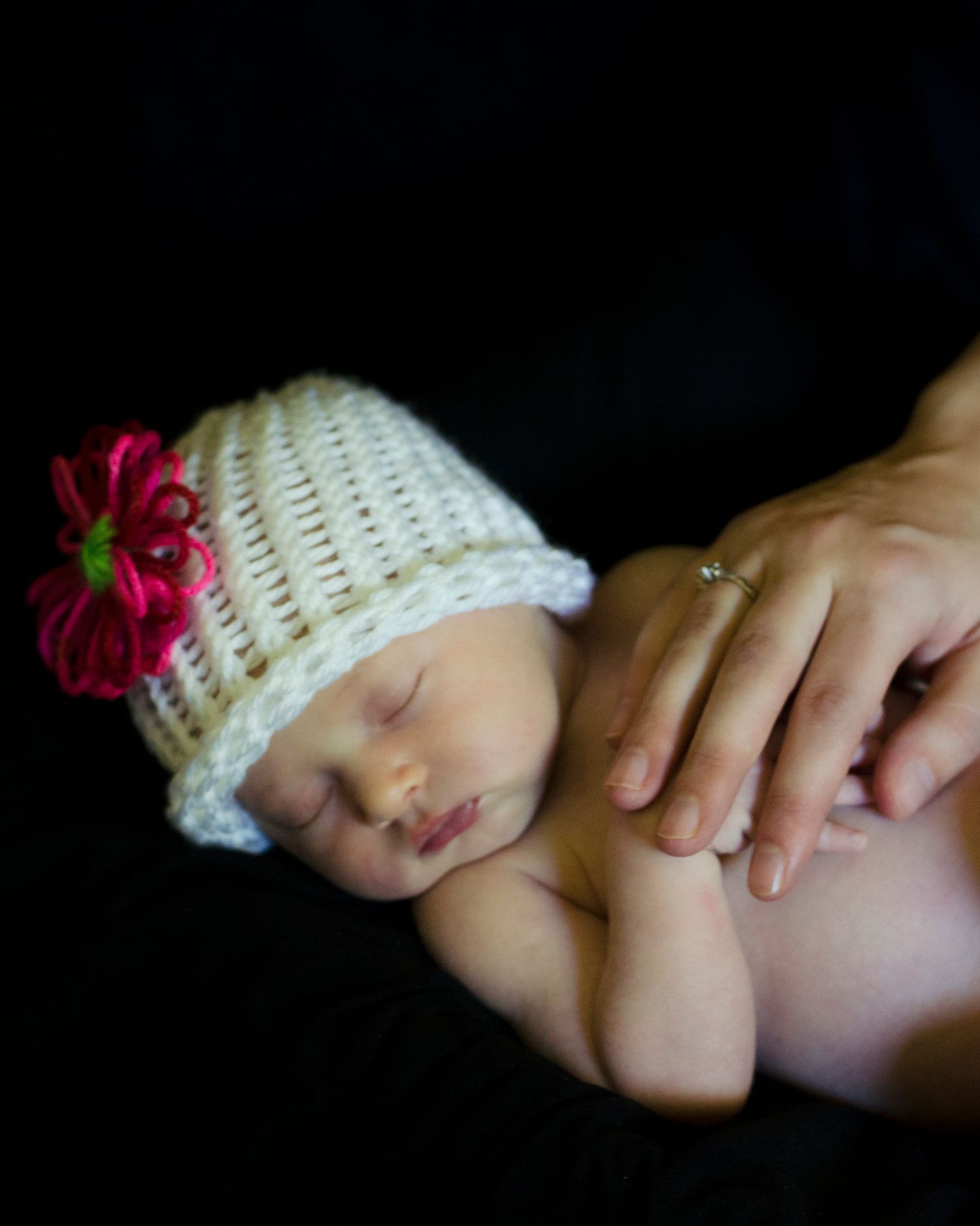 A sleeping baby wearing a white knit hat with pink flower detail rests against a black background.