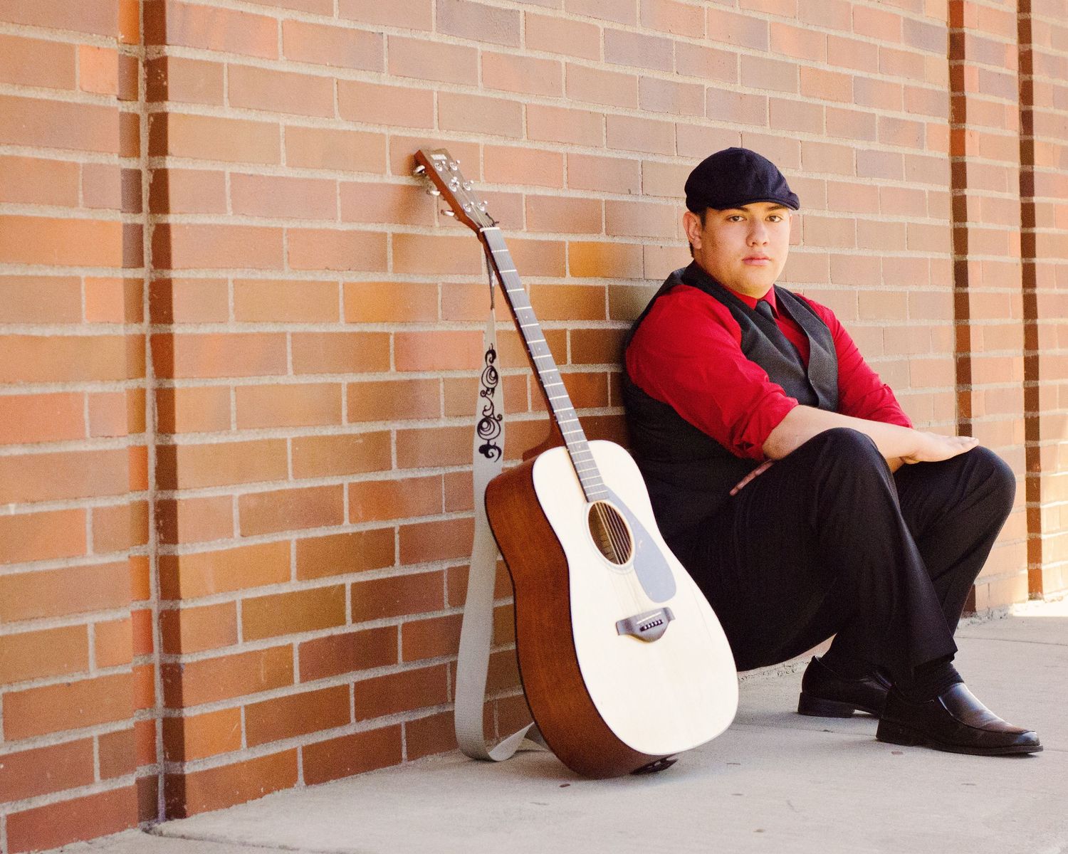 A person sits on wooden flooring against a brick wall while holding an acoustic guitar.