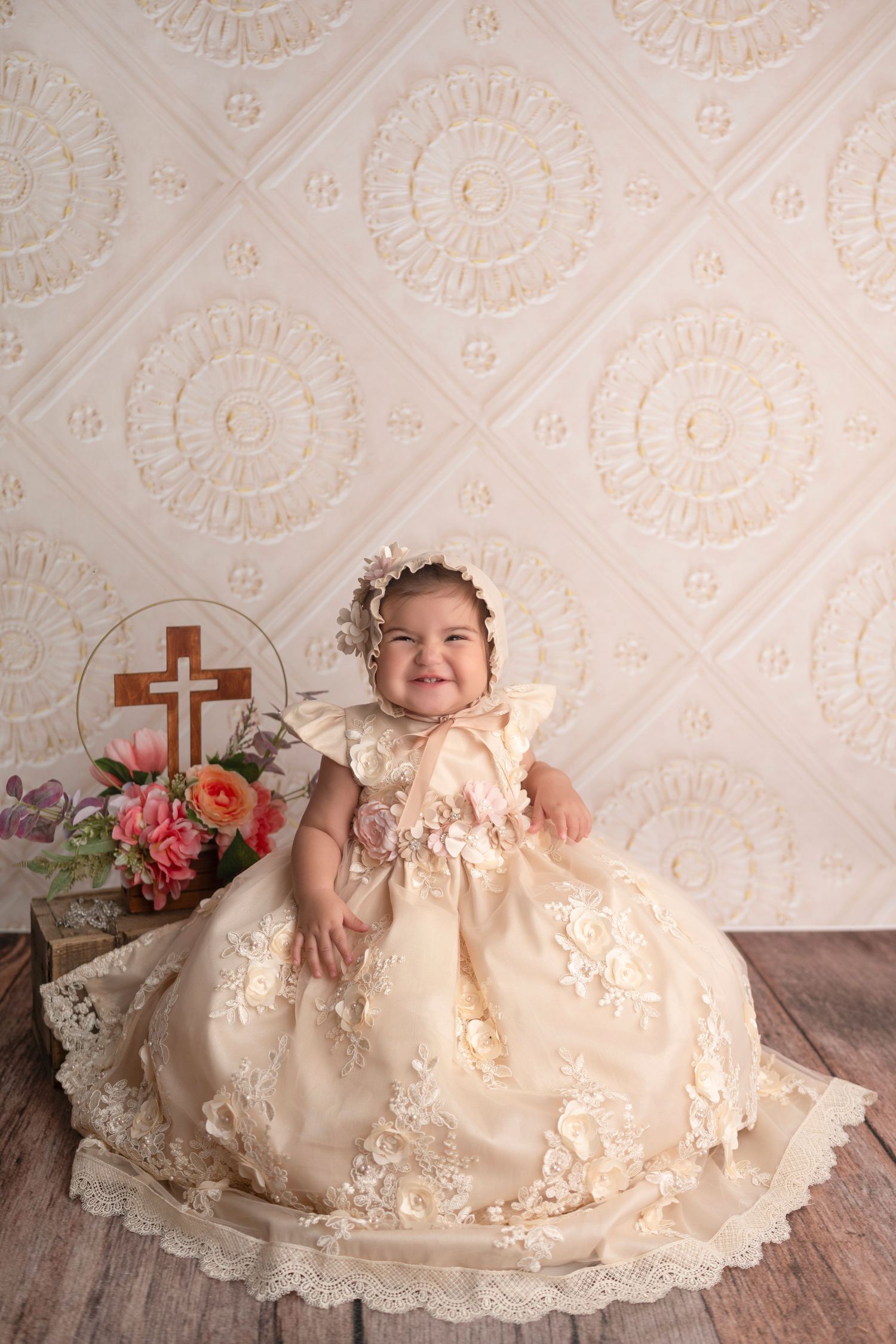 Baby in ornate cream baptismal gown sitting by cross and flowers against textured backdrop.