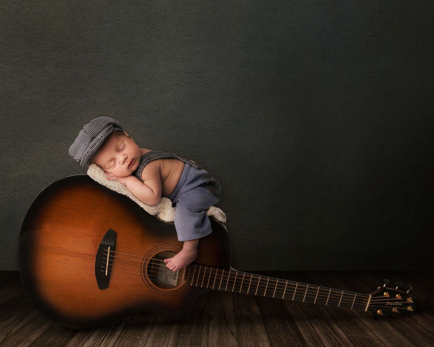 A sleeping newborn in grey outfit rests peacefully on a large brown acoustic guitar against dark backdrop.