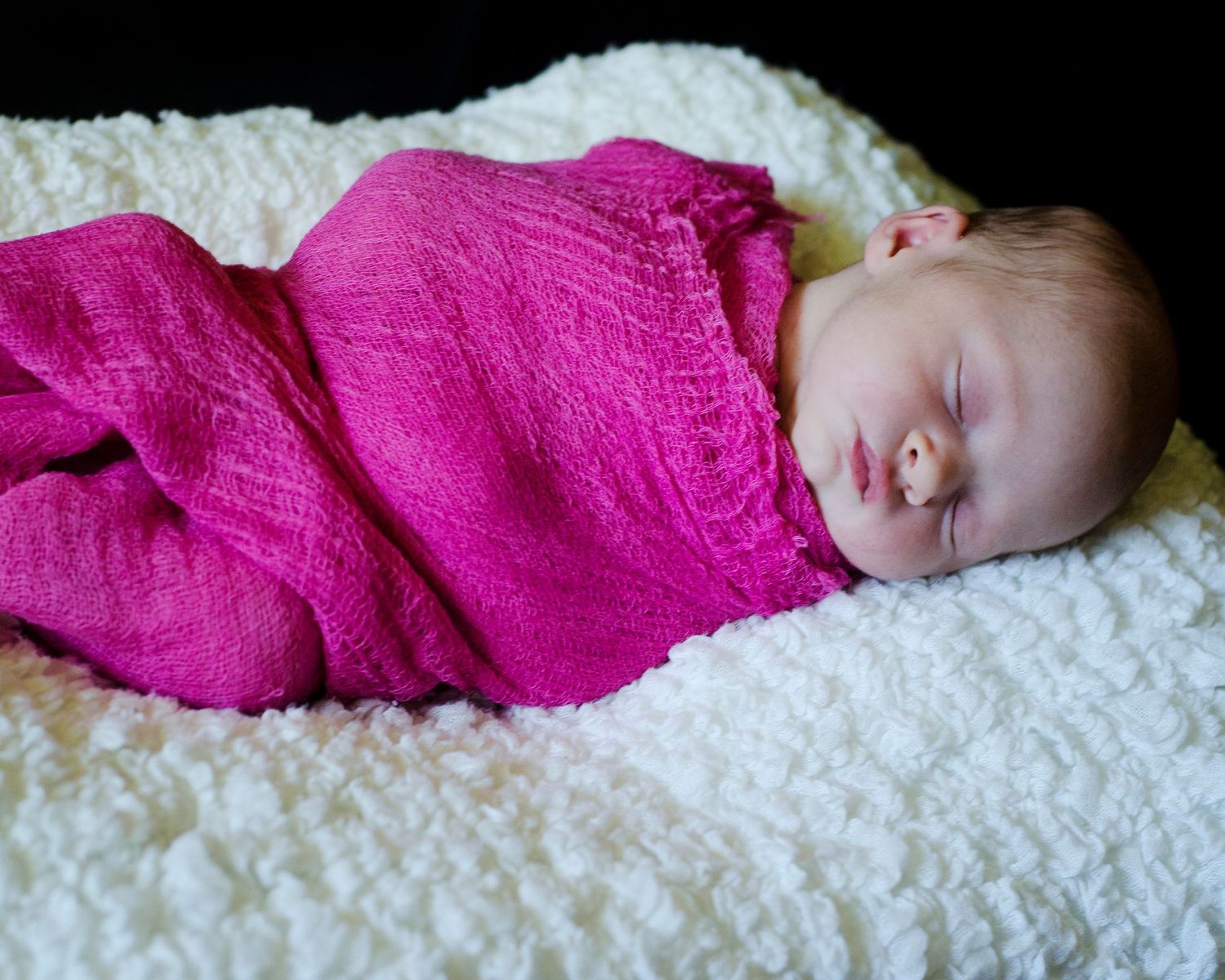 Sleeping baby peacefully wrapped in bright pink blanket on soft white textured surface.