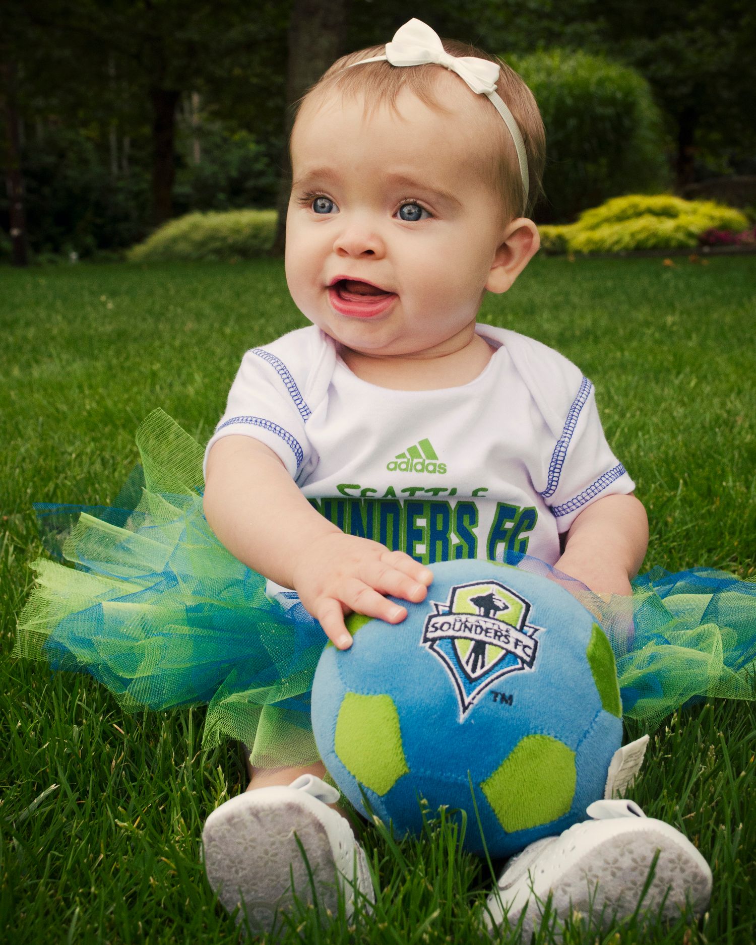 Baby wearing white Adidas outfit sits on grass with blue and green Seattle Sounders soccer ball.