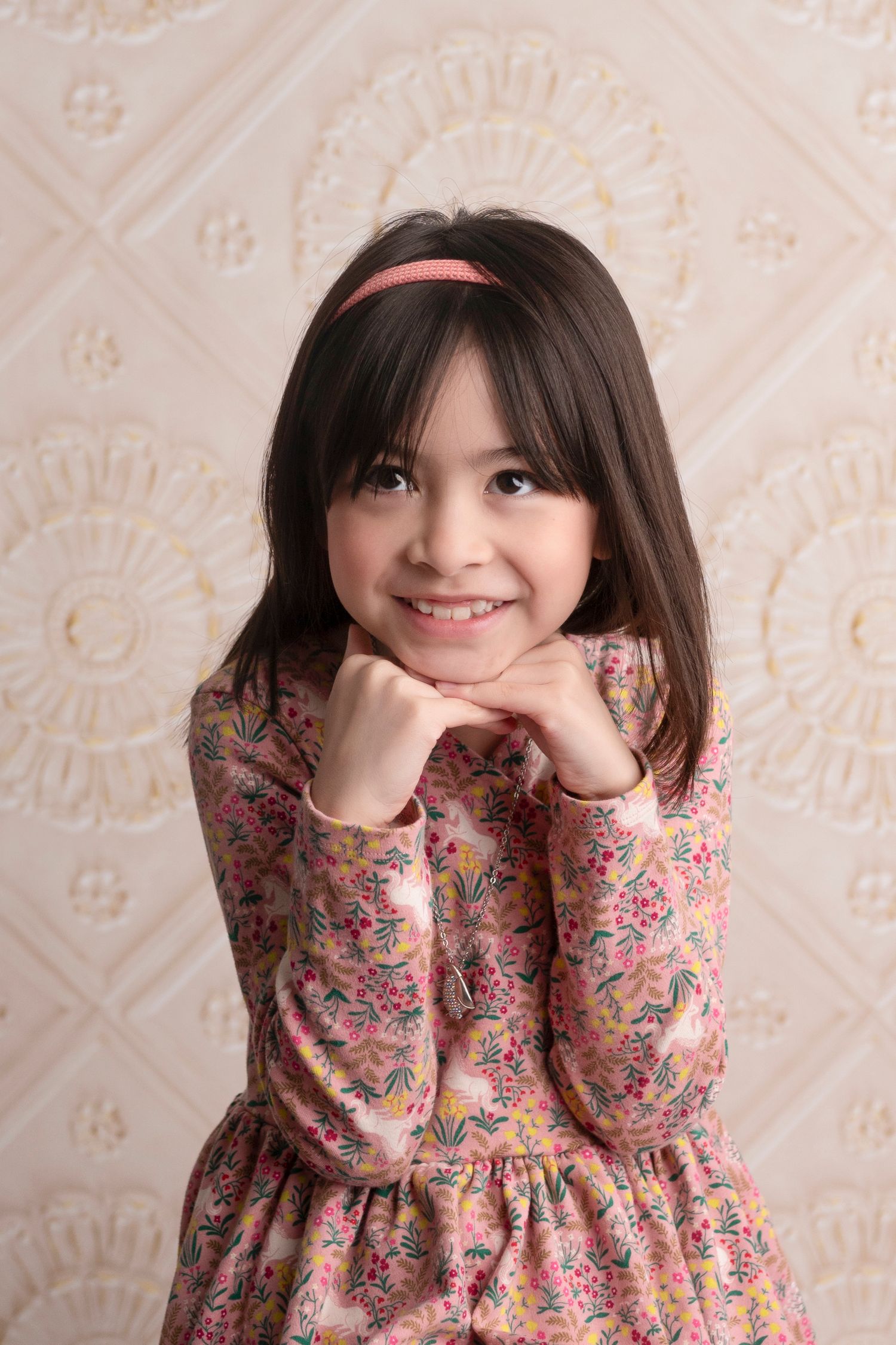 Young girl in floral dress with bangs smiling at camera against textured background.