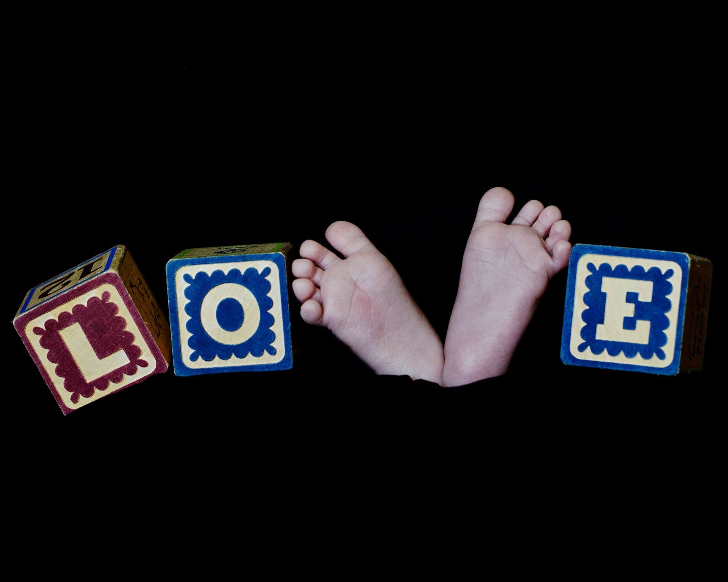 Newborn baby feet between colorful alphabet blocks spelling out LOVE against black background.