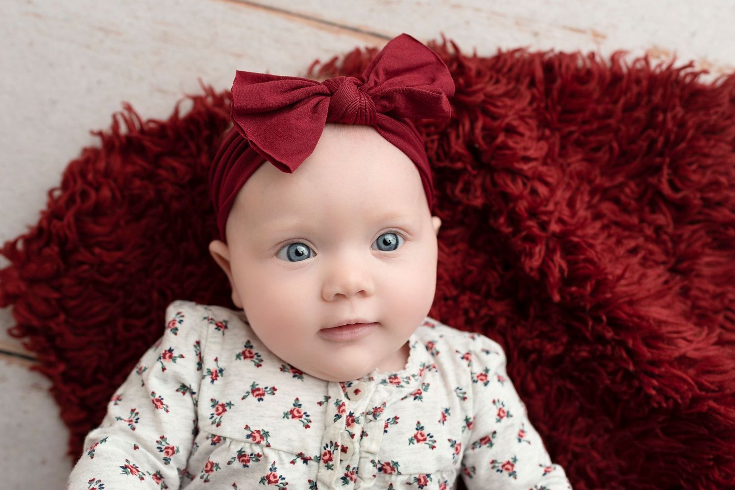 Baby wearing floral outfit with red bow lying on crimson blanket looking at camera.
