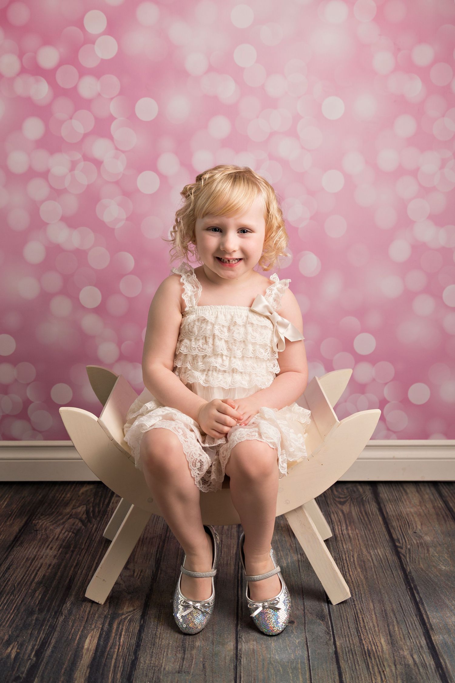 Small child sits in white chair wearing ruffled pink dress and silver shoes against pink bokeh backdrop on wooden floor.