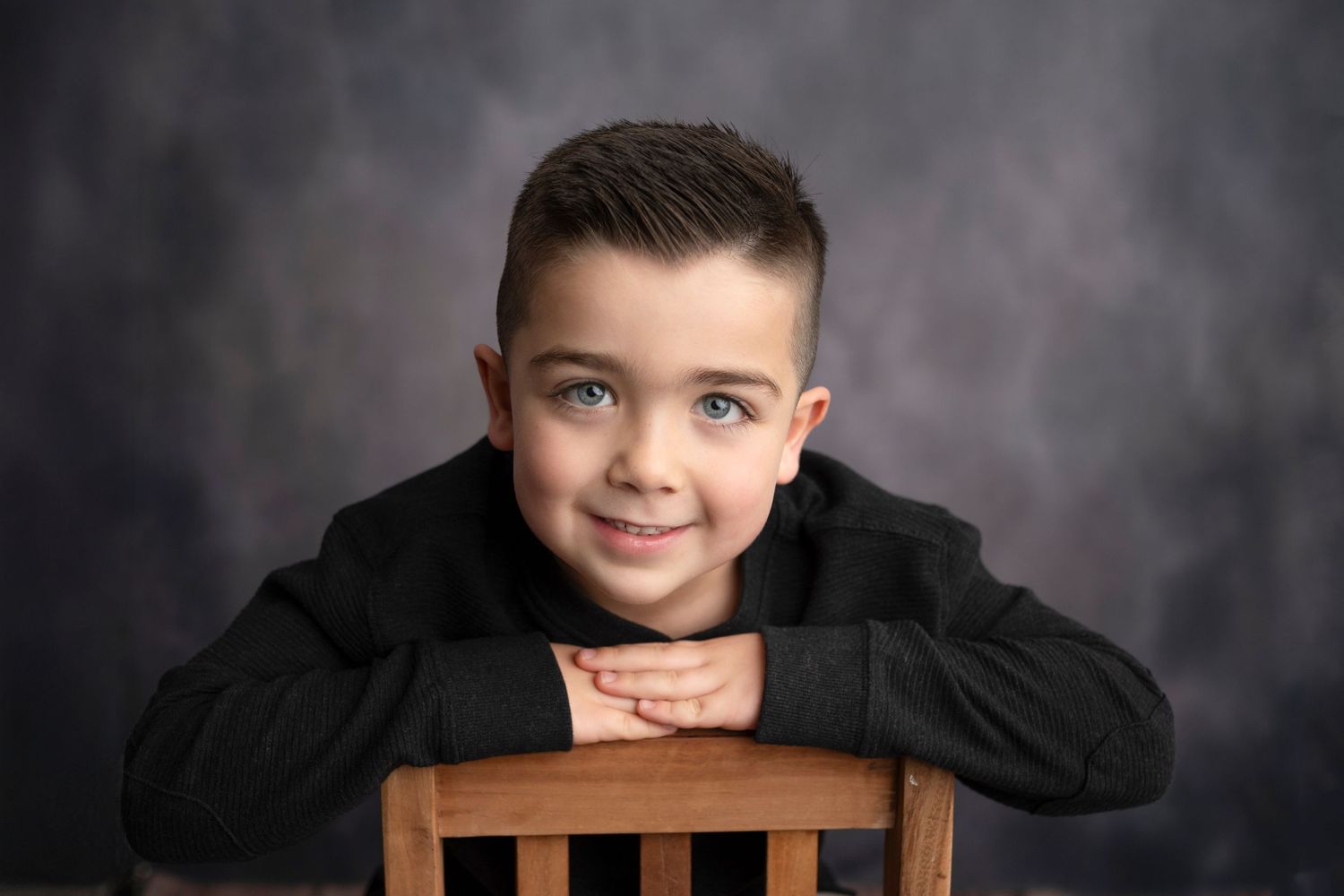 Portrait session featuring child in black sweater posing with wooden chair against grey backdrop.